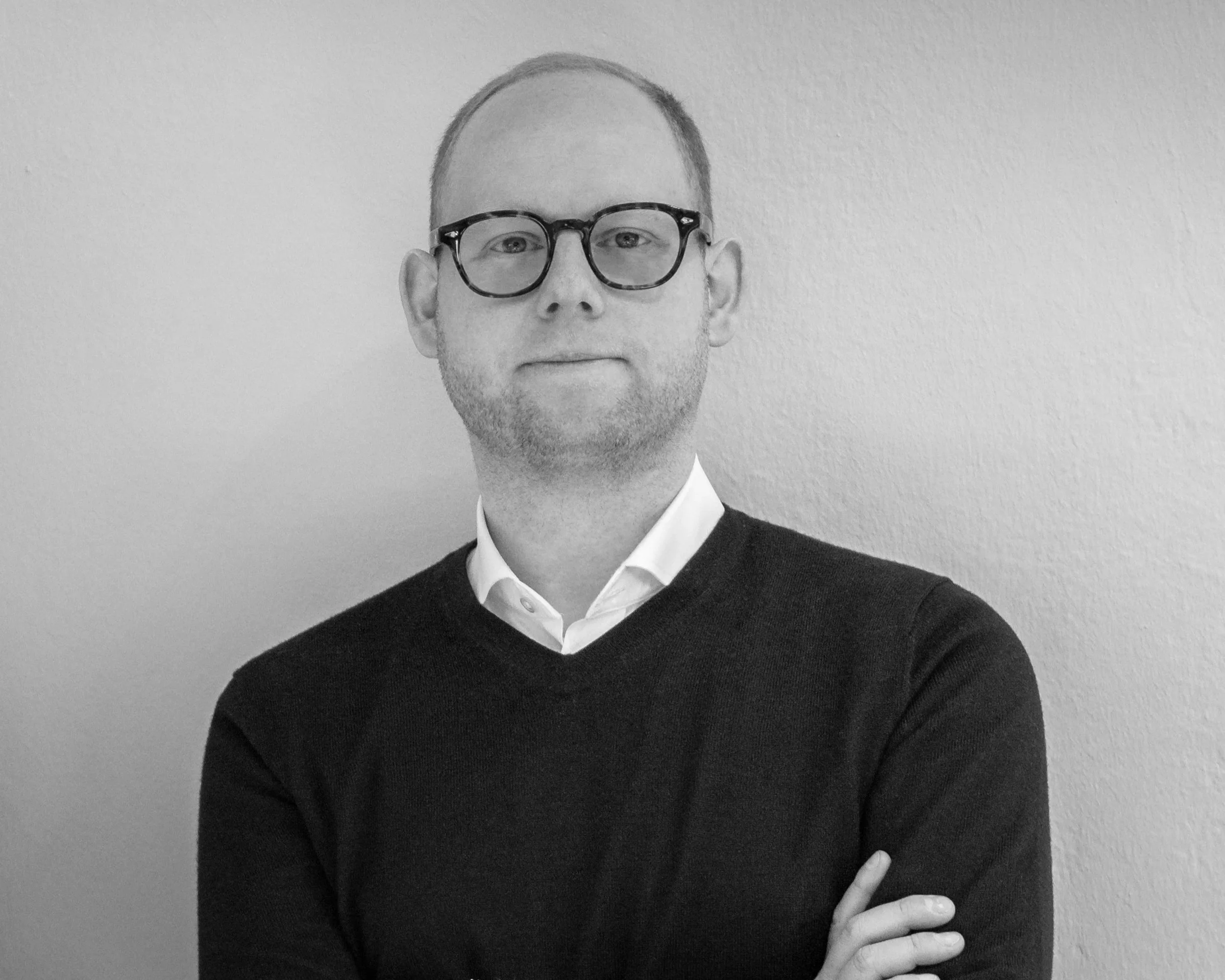Schwarz-weiß-Porträt eines Mannes mit Brille, weißen Hemd und schwarzem Pullover, vor einer hellen Wandstehend mit verschränkten Armen.
