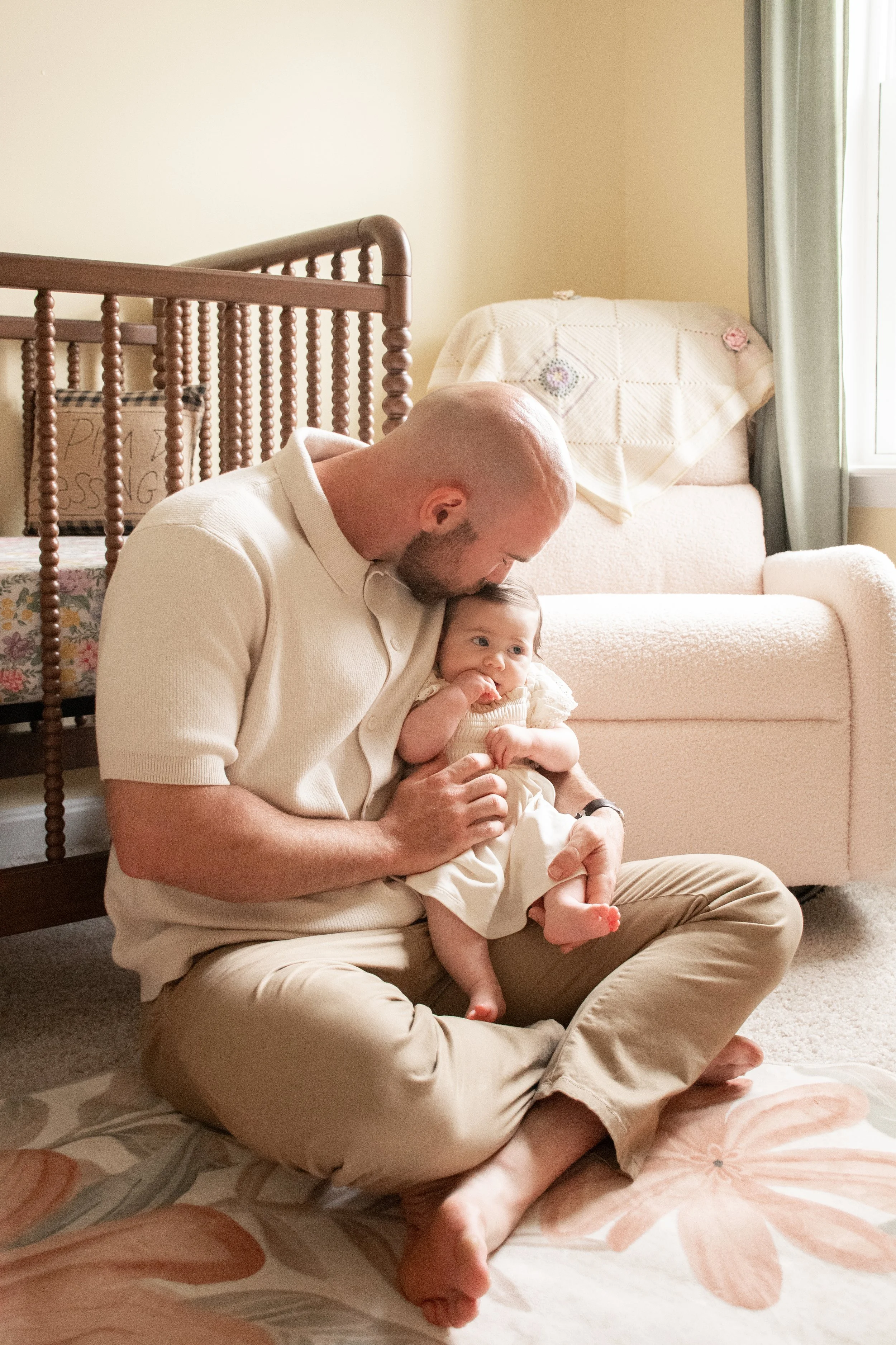 A father sitting cross-legged on the floor holding his baby daughter in a cream dress.
