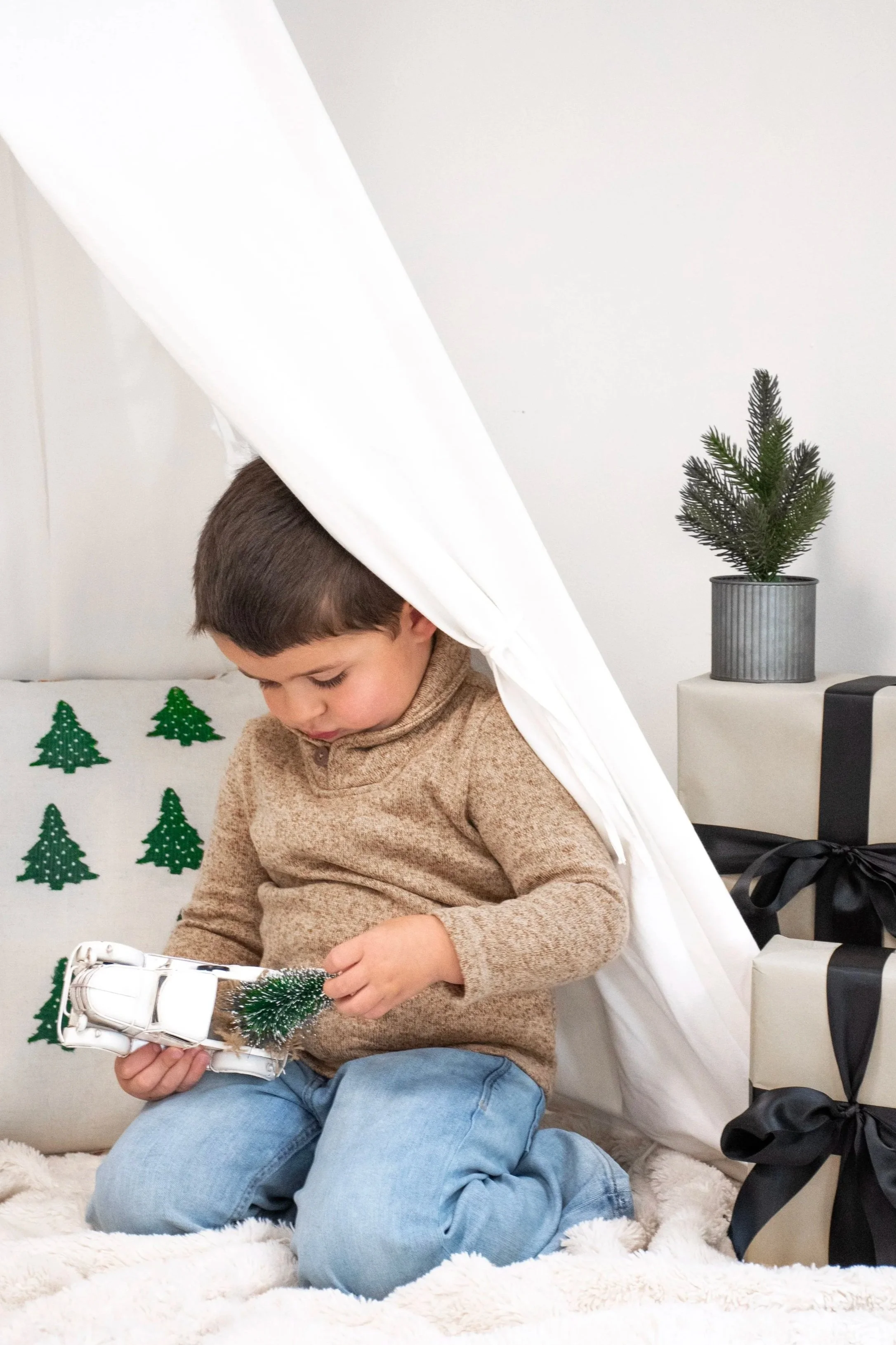 A young boy sitting on a soft, furry rug under a white cloth tent, holding a small Christmas tree ornament and a tiny miniature Christmas tree. The background includes a pillow with green Christmas trees and a festive wrapped gift with black ribbon, along with a small potted pine tree.