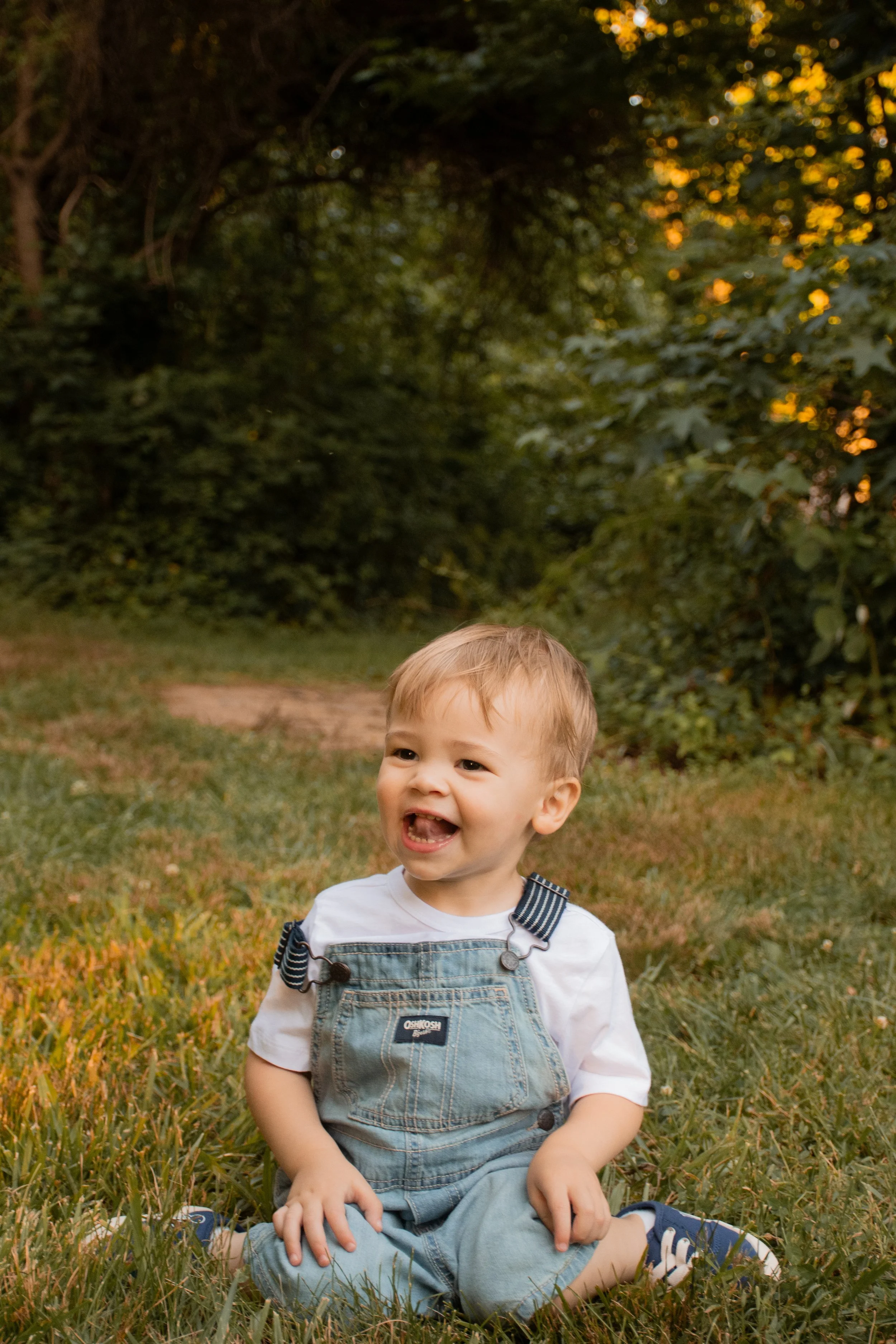 A toddler in denim overalls sitting in the grass, smiling