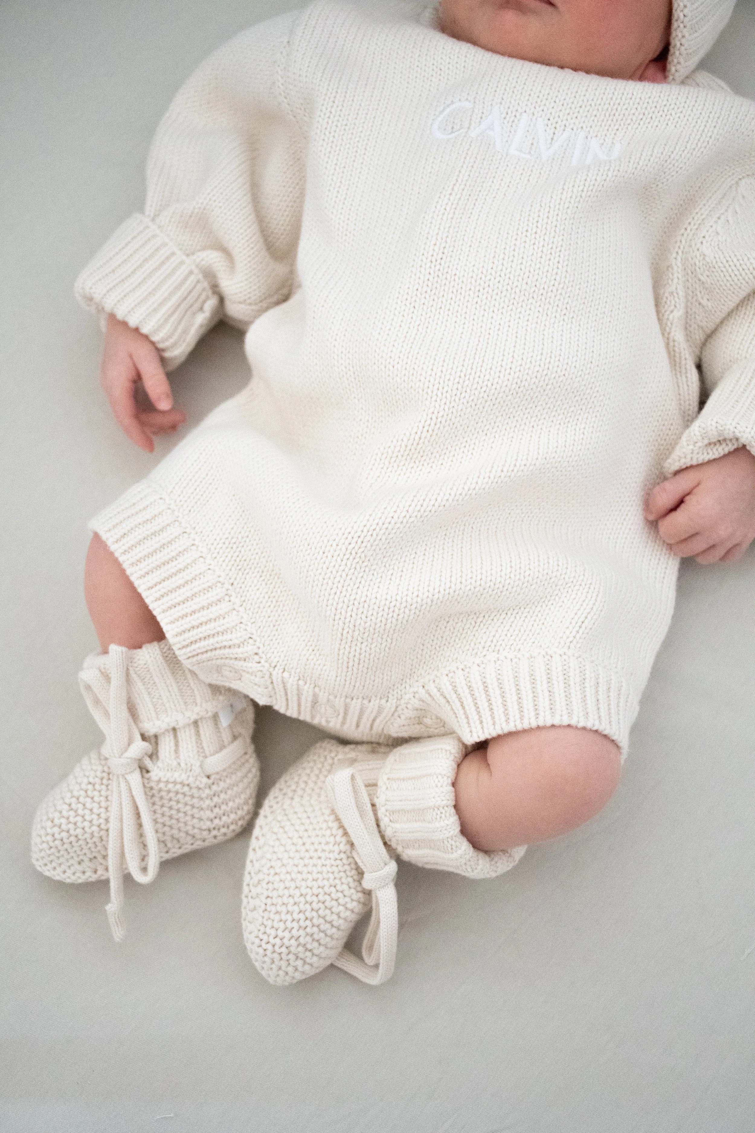 A newborn in a cream knit outfit and booties lying on a soft blanket