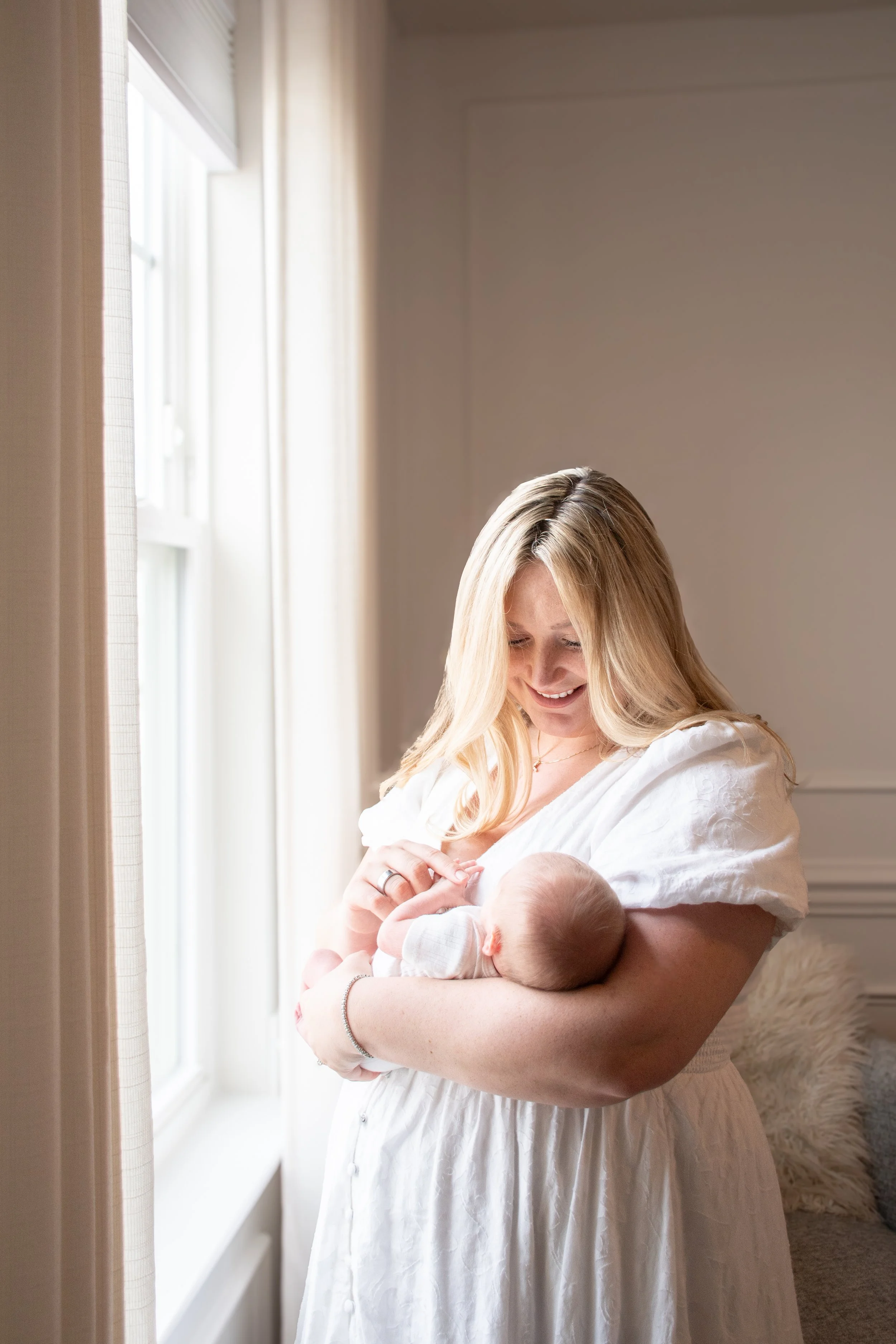 A mother in a white dress holding her newborn by a window