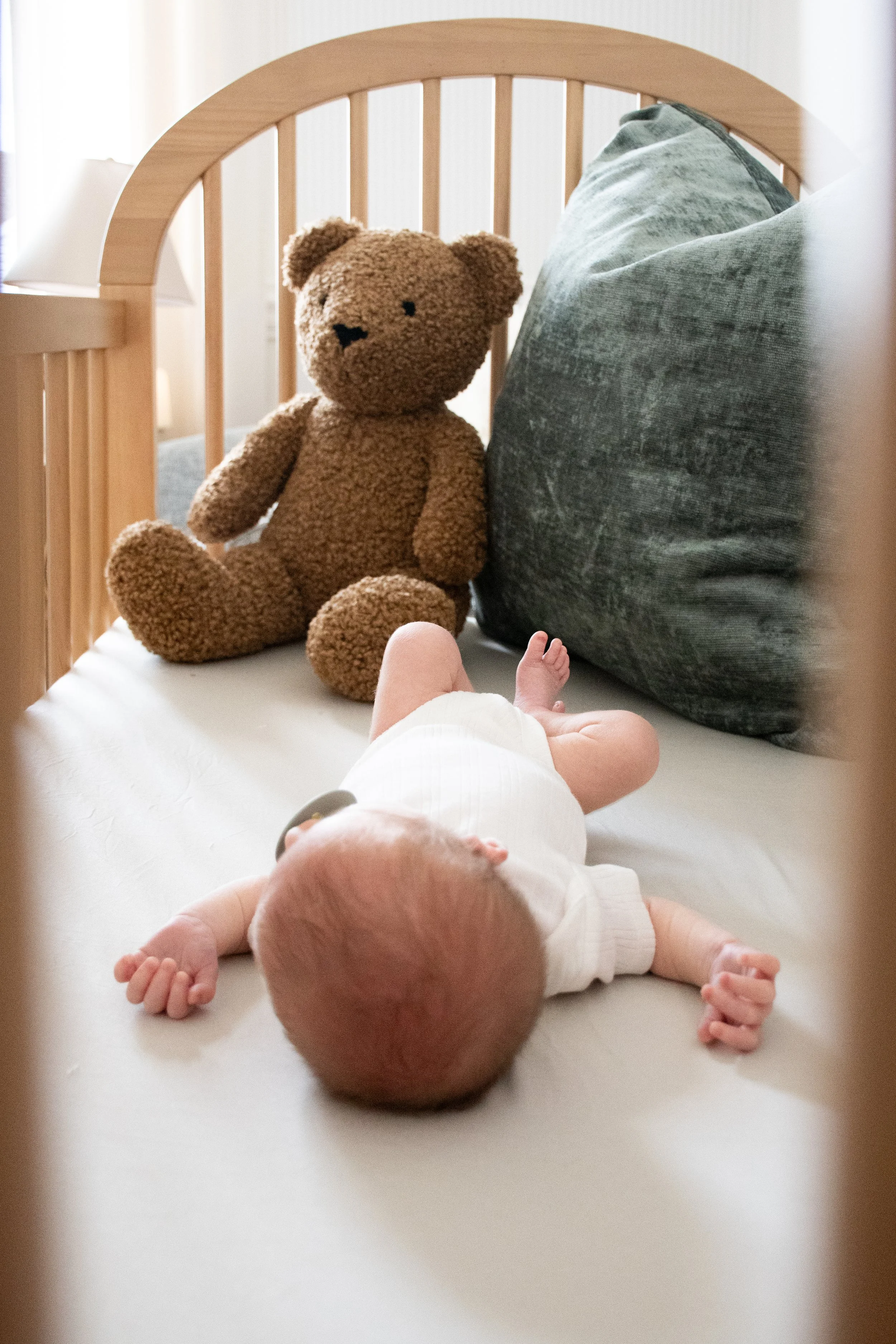 A newborn lying on a bed beside a teddy bear, a wooden crib behind