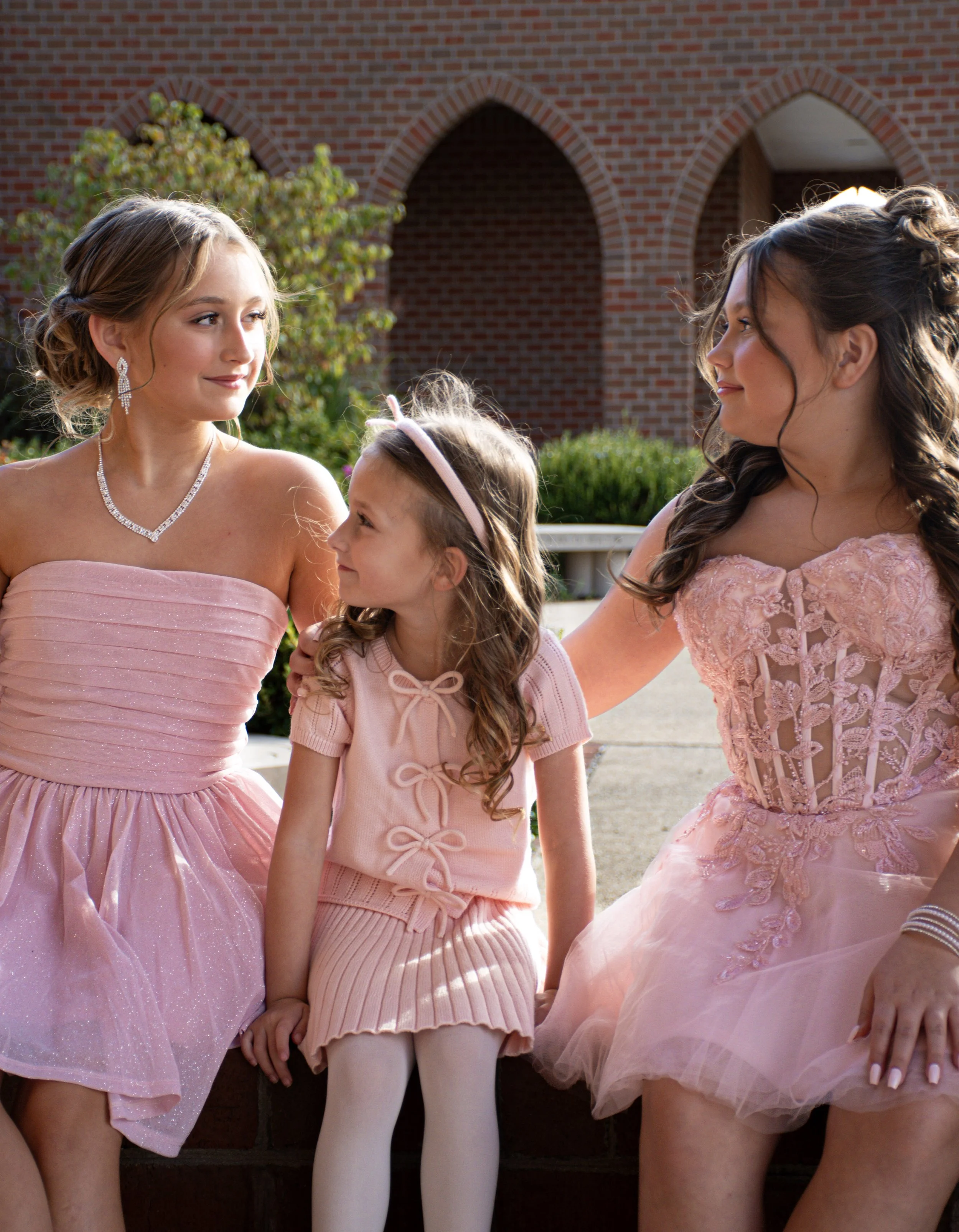 Three females, two young women and one girl, dressed in pink dresses, sit outdoors in front of a brick building and green shrubs, smiling and engaging with each other.