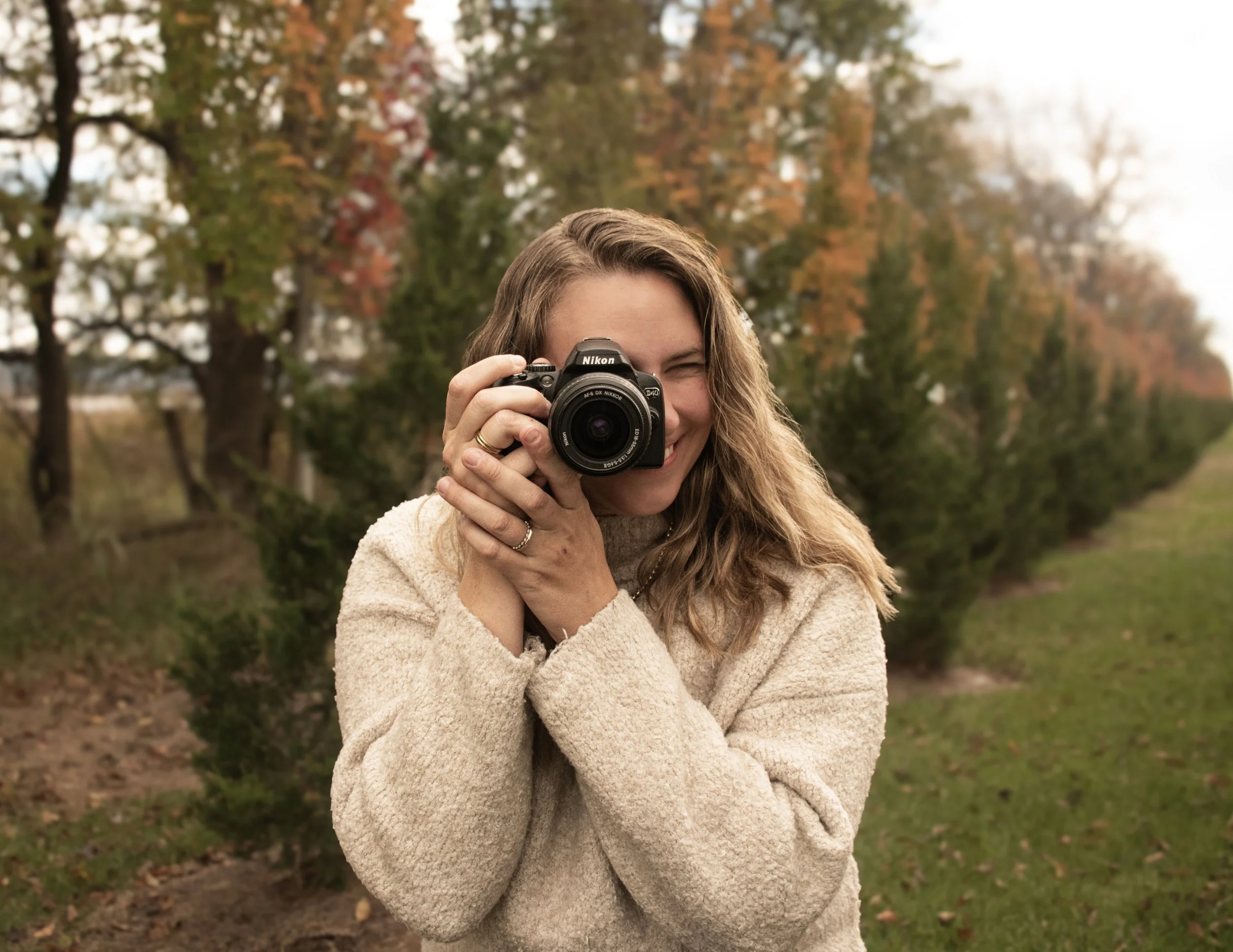 A woman smiling outdoors holding a professional camera up to her face, with fall foliage in the background.