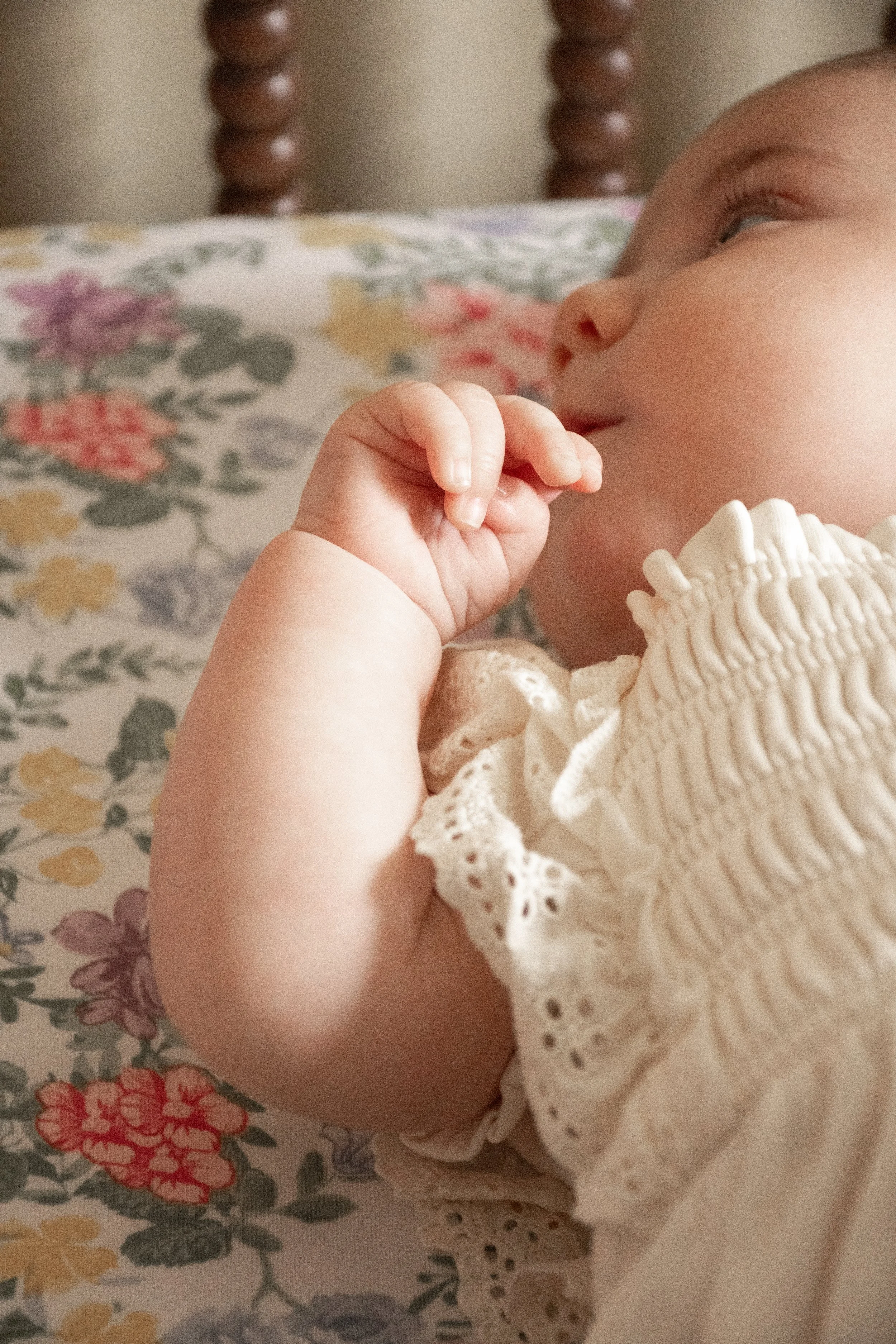 A close-up of a baby in a cream textured dress resting on a floral surface, fingers near her mouth.