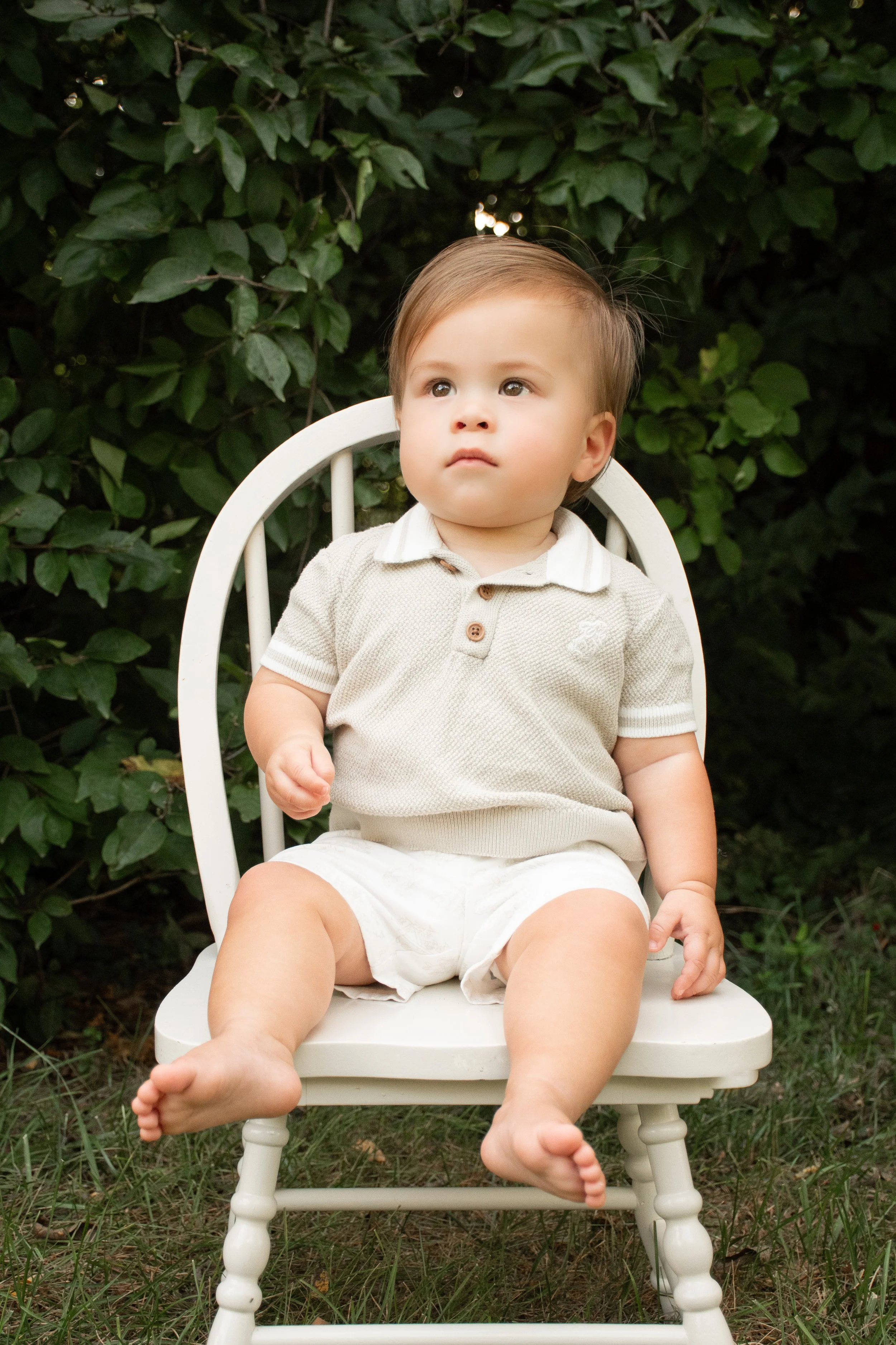 A young boy sitting in a white, wooden chair in front of a wall of greenery.