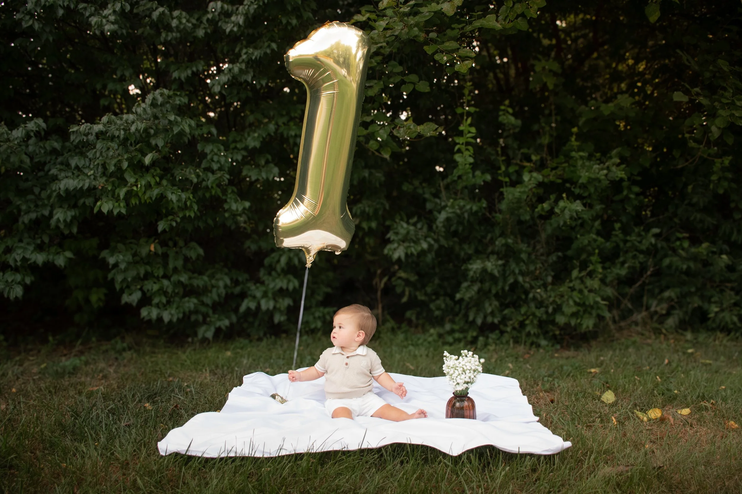 A young boy sitting on a white blanket in a grassy field with a vase of baby's breath flowers and a floating #1 golden ballon.
