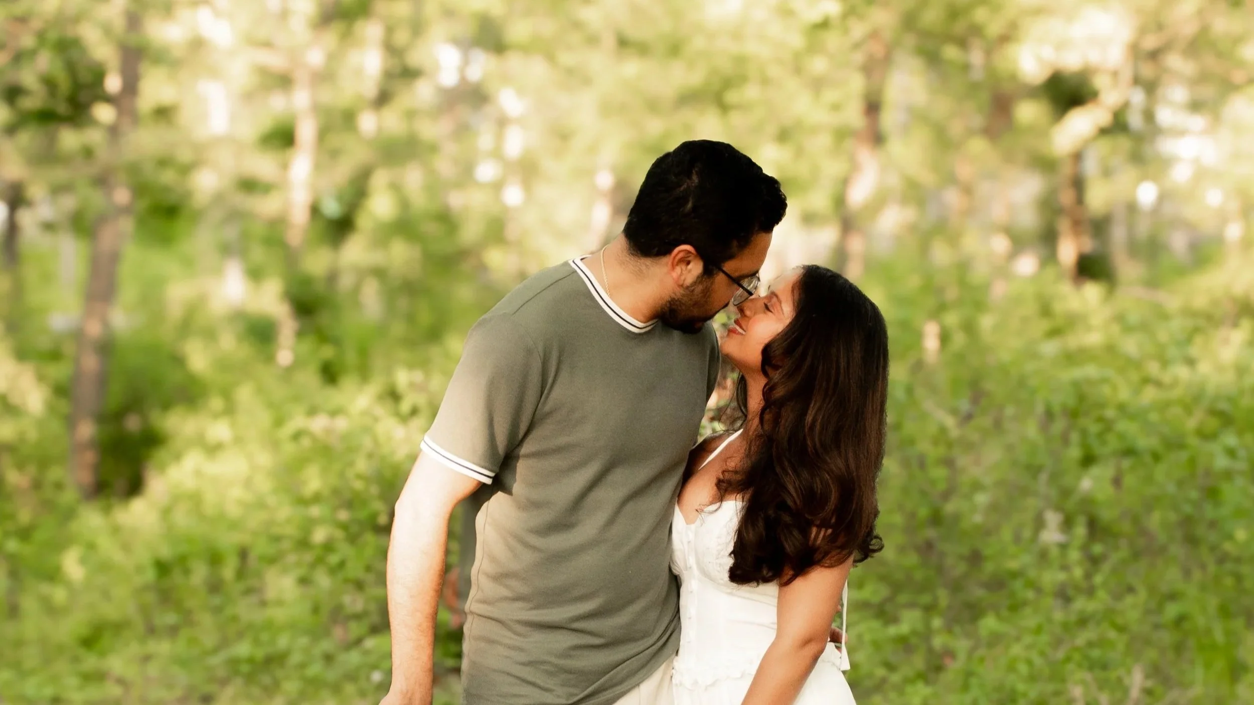 A couple standing in a wooded area leaning in to kiss each other in the summertime.