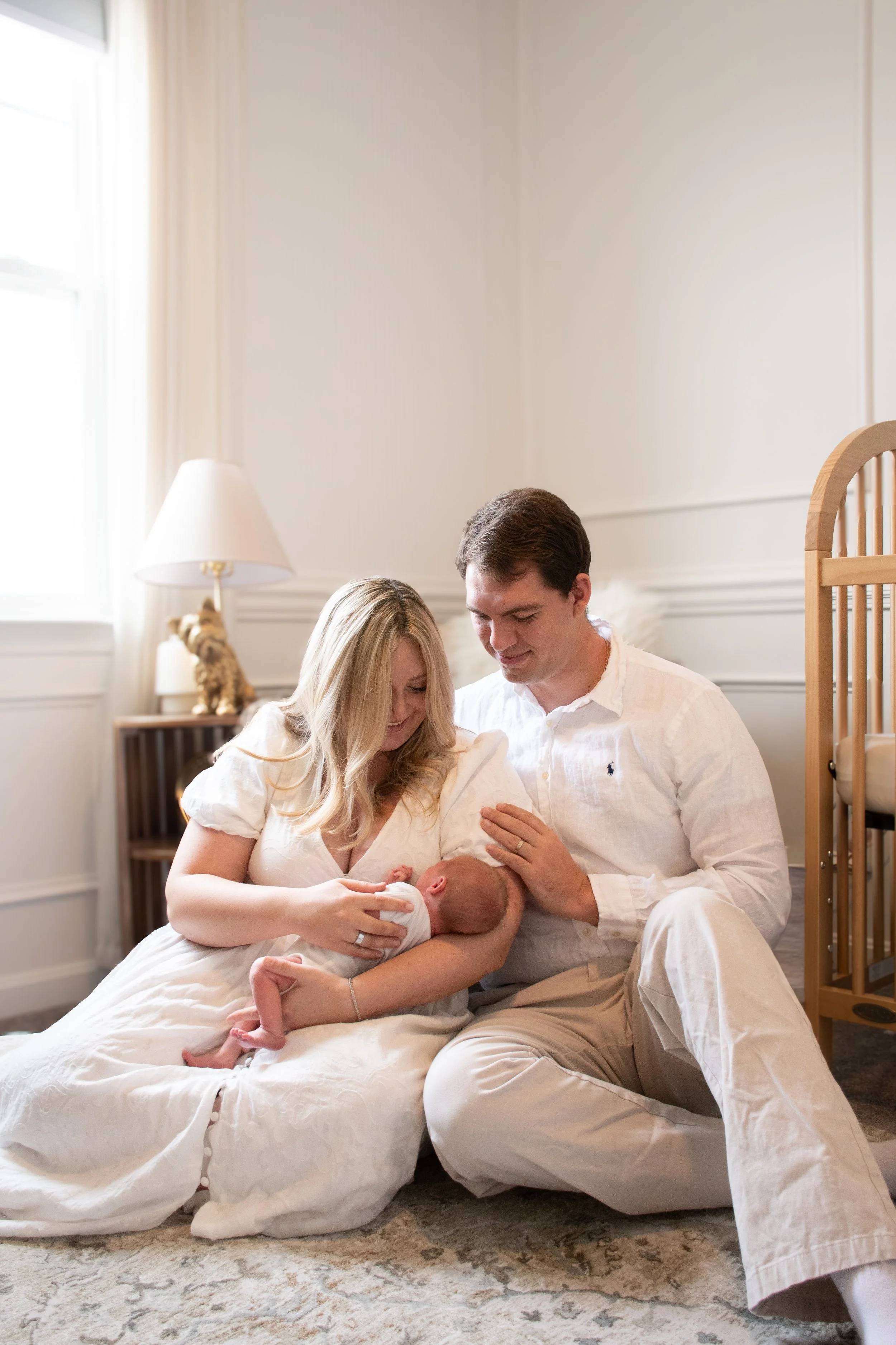 Parents sitting on the floor, the mother holding their newborn as both smile down at the baby.