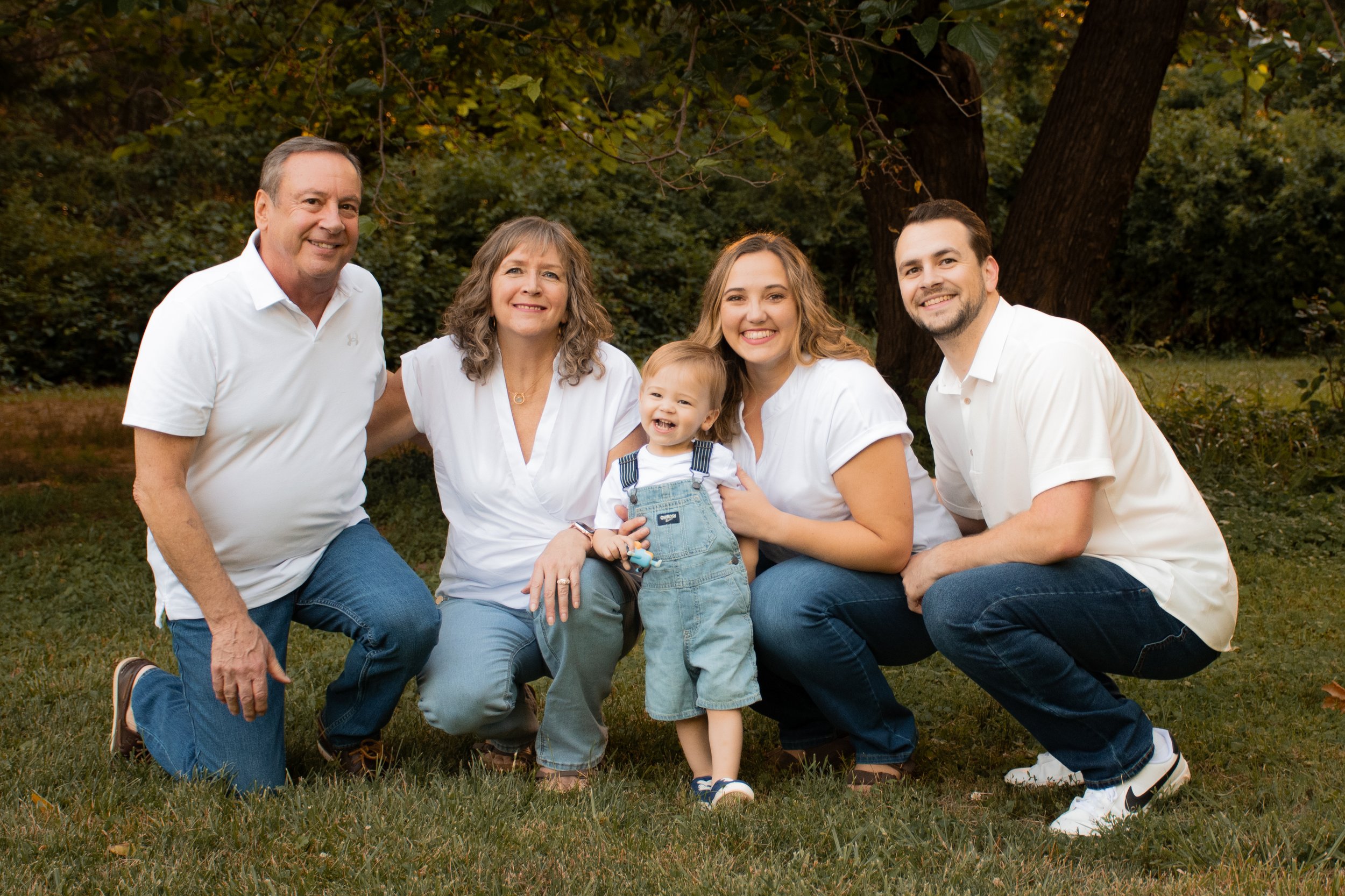 A family of five kneeling in a grassy field with green foliage in the background in the summertime.