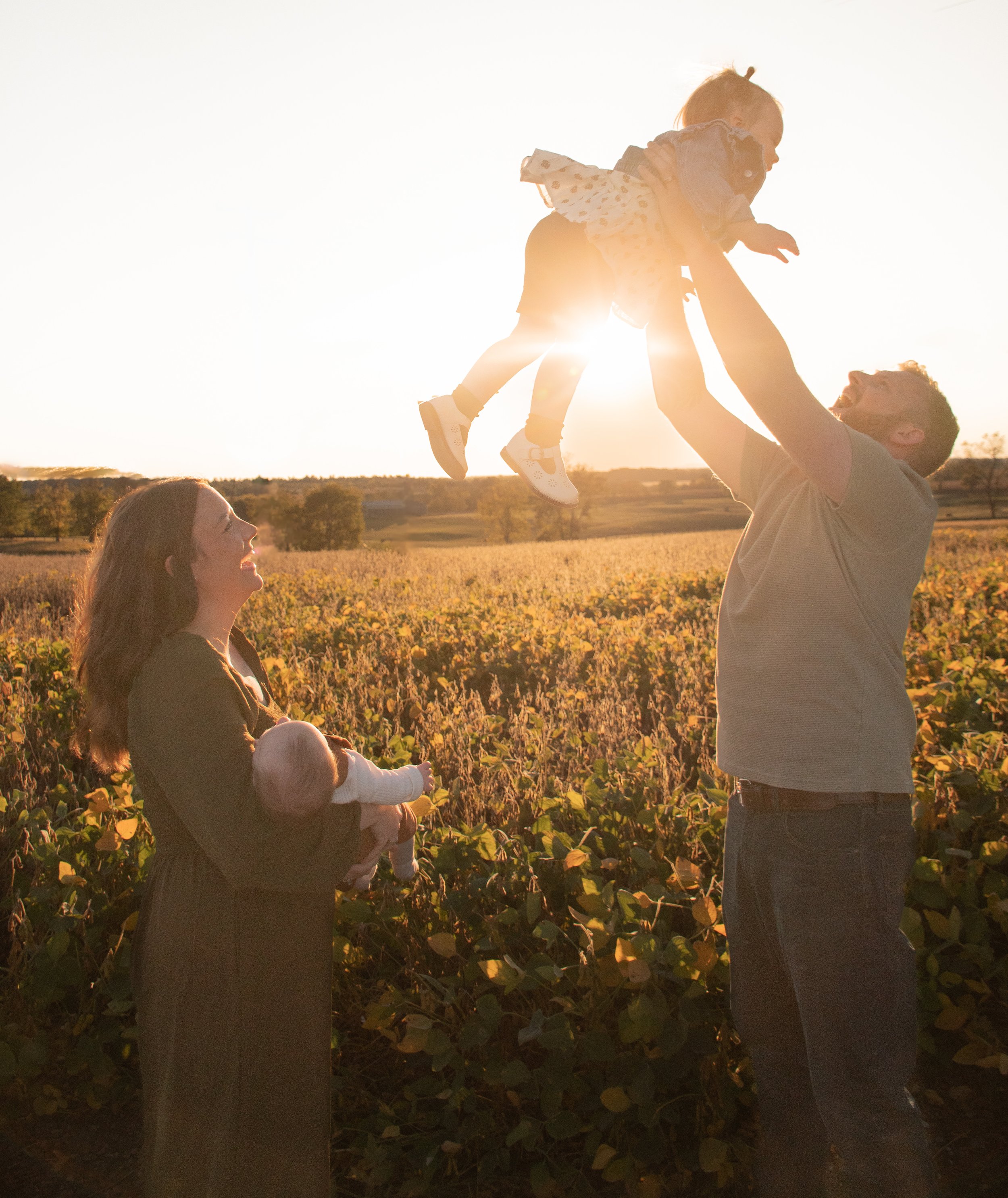 A family of four standing in a field at golden hour, the mom holding an infant and looking up at the dad throwing a toddler girl in the air