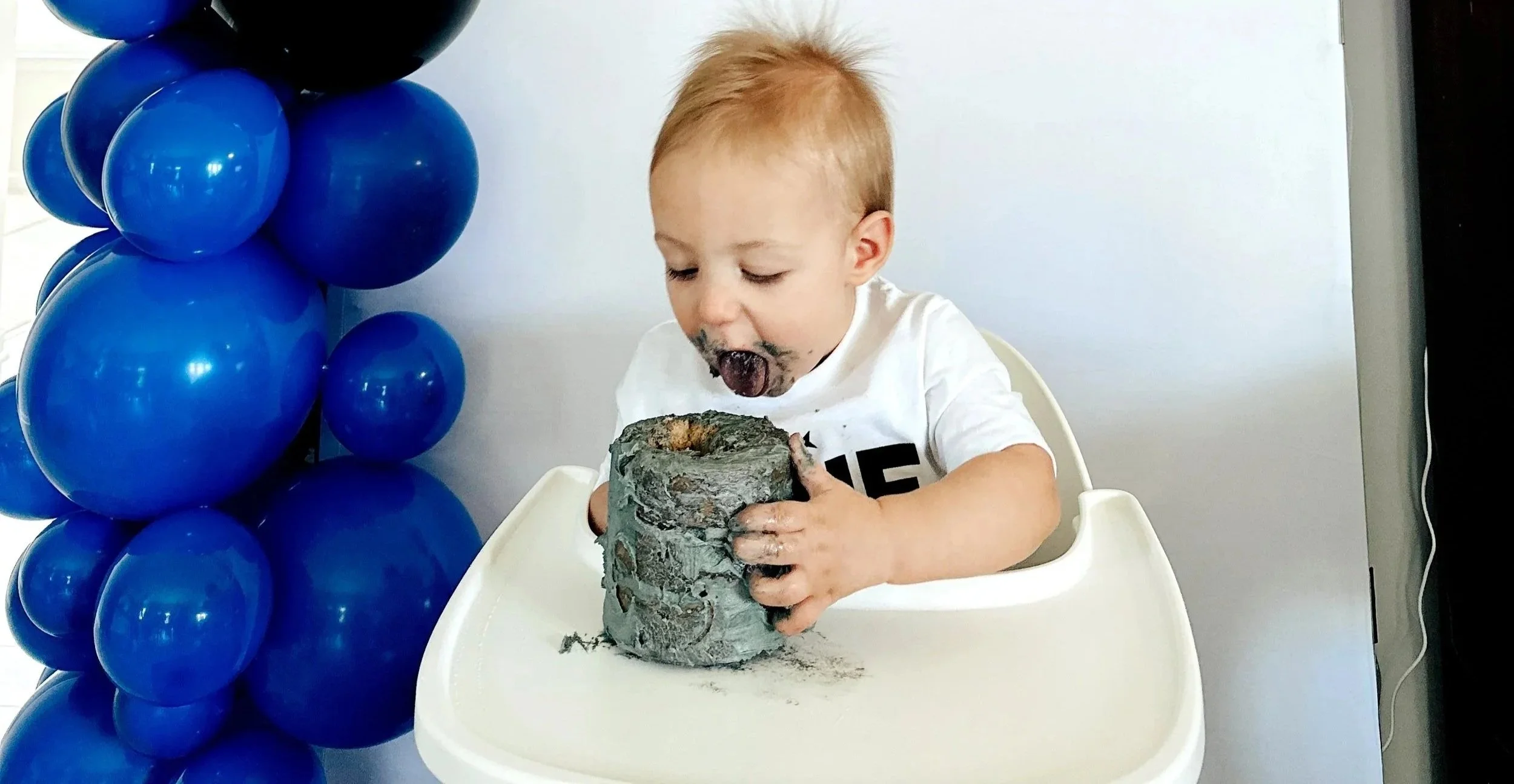 A one year old child in a highchair, diving into a birthday cake at his space themed birthday party with cake all over his face.