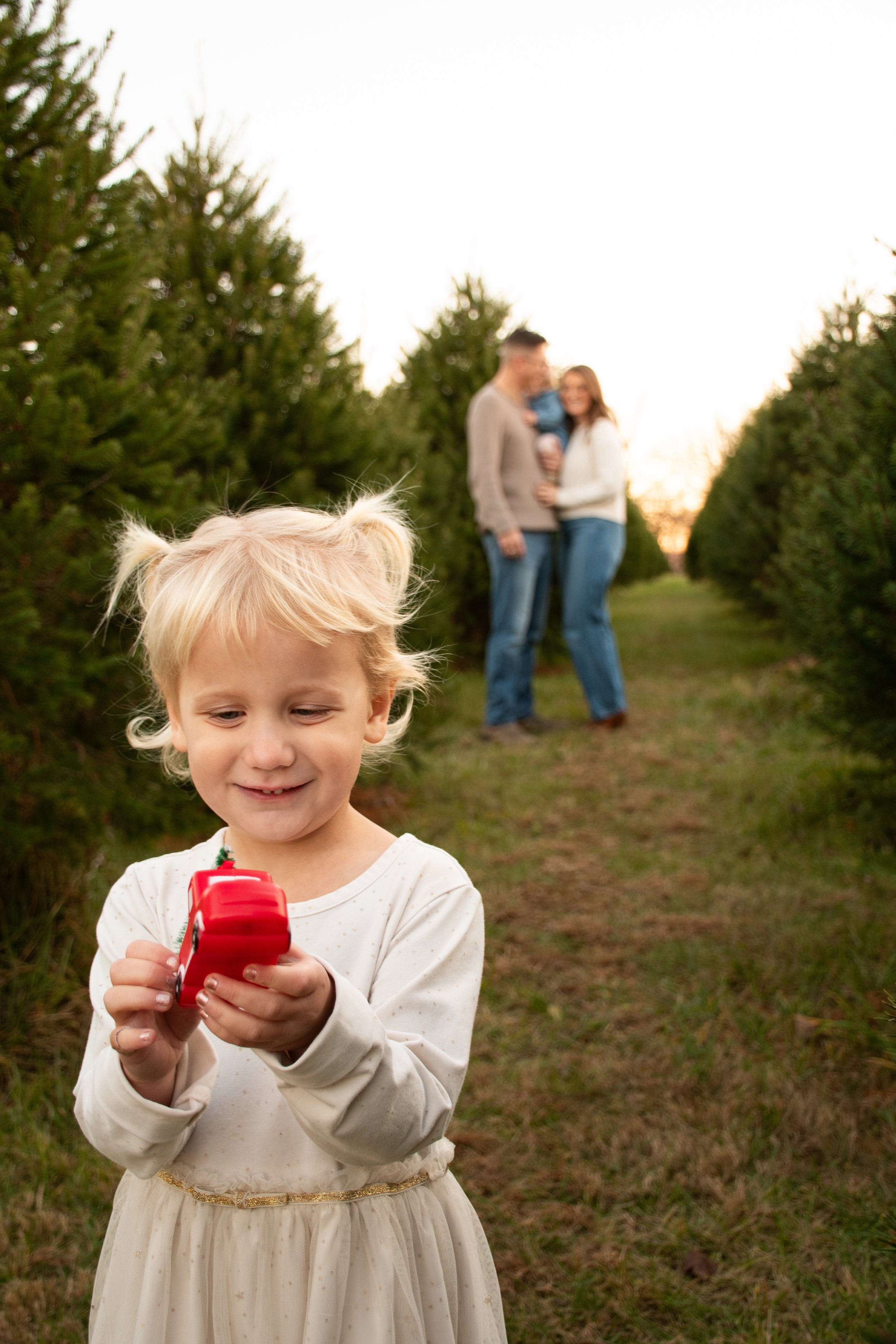 A girl in pigtails smiles at a red toy in her hands, her family blurred behind her at sunset.