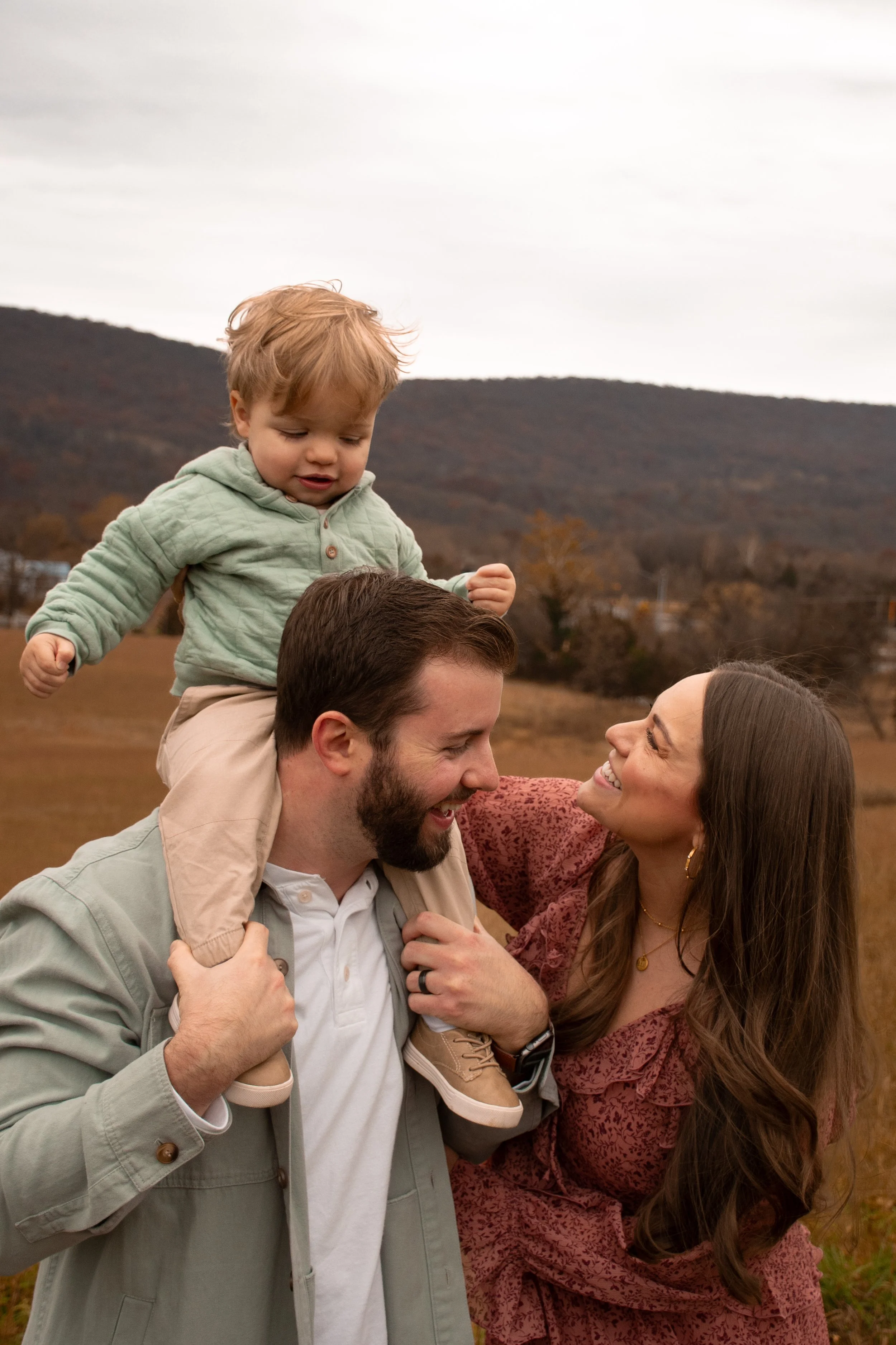 A father carries his son on his shoulders while the mother smiles at them, in an open field below distant hills.