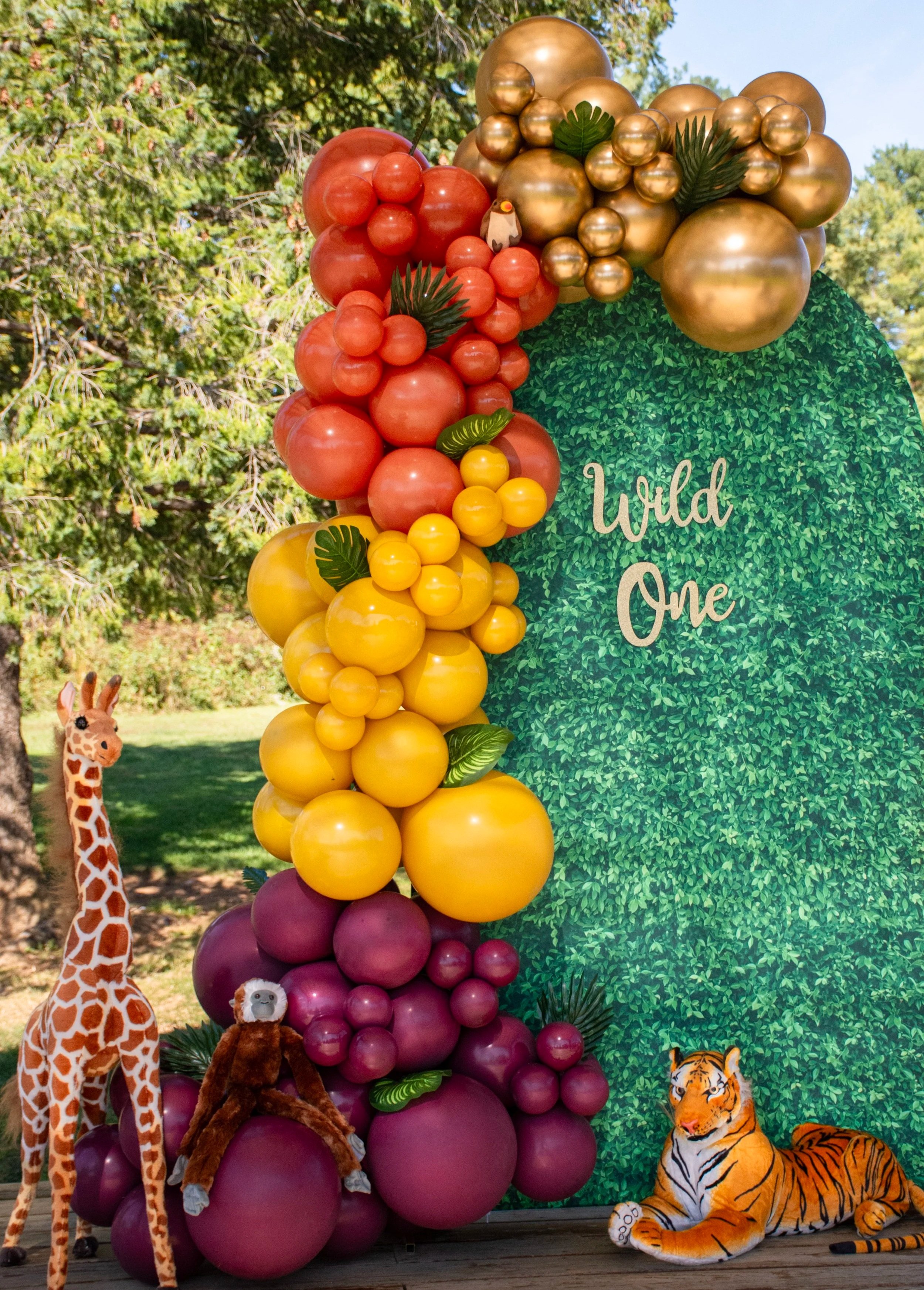 Colorful balloon display with a green leafy background and the words "Wild One" on it, featuring stuffed animals including a giraffe, a monkey, and a tiger, outdoor setting with trees.