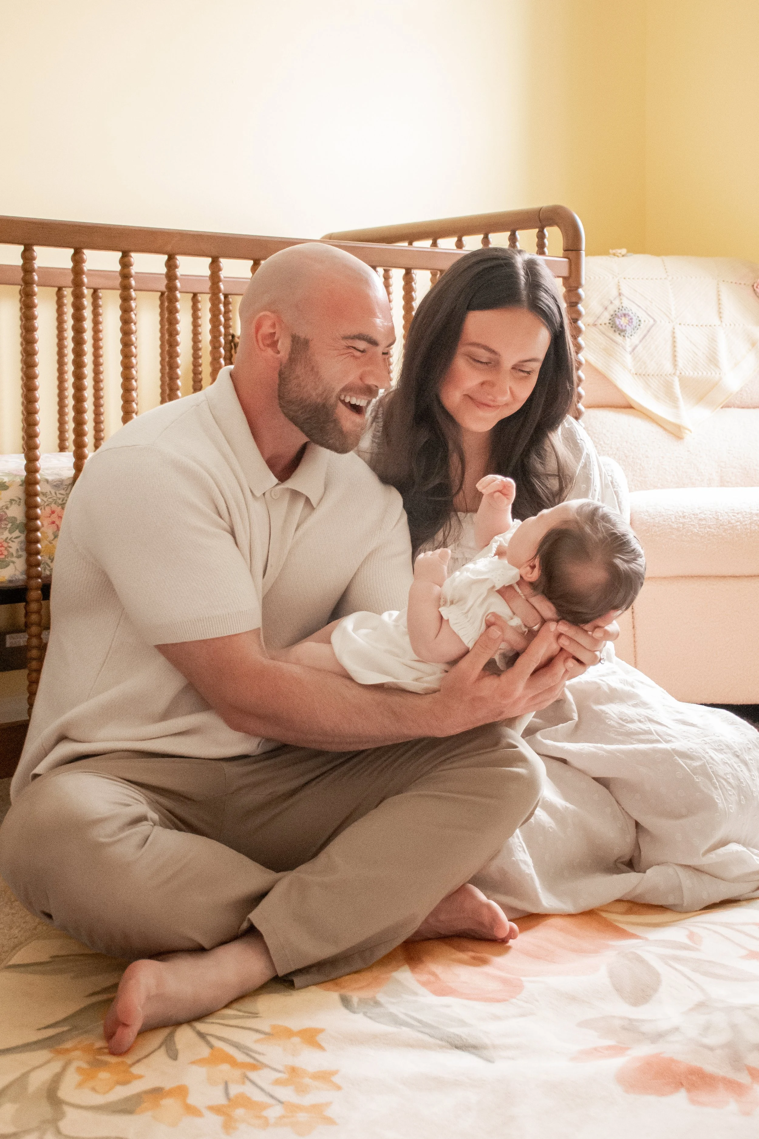 Parents sitting on the floor in front of a crib, holding their baby and smiling down at them.