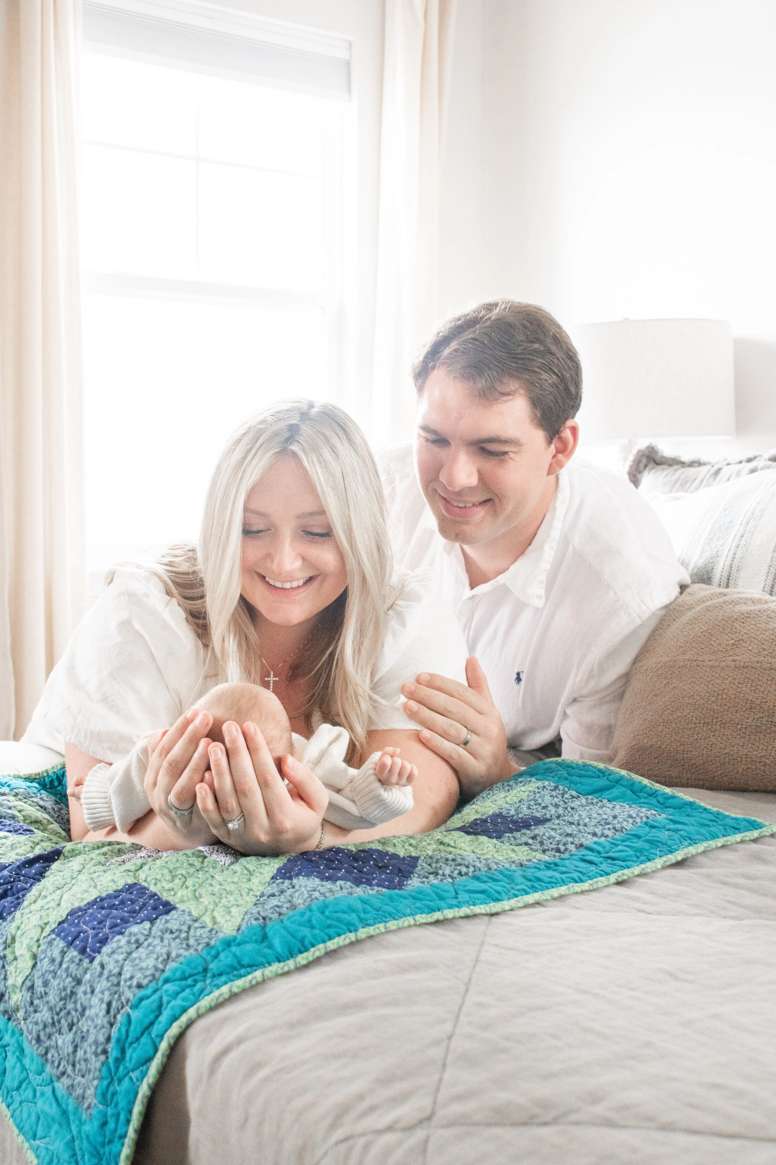 A mother holding her newborn on a quilted bed while the father smiles behind her.
