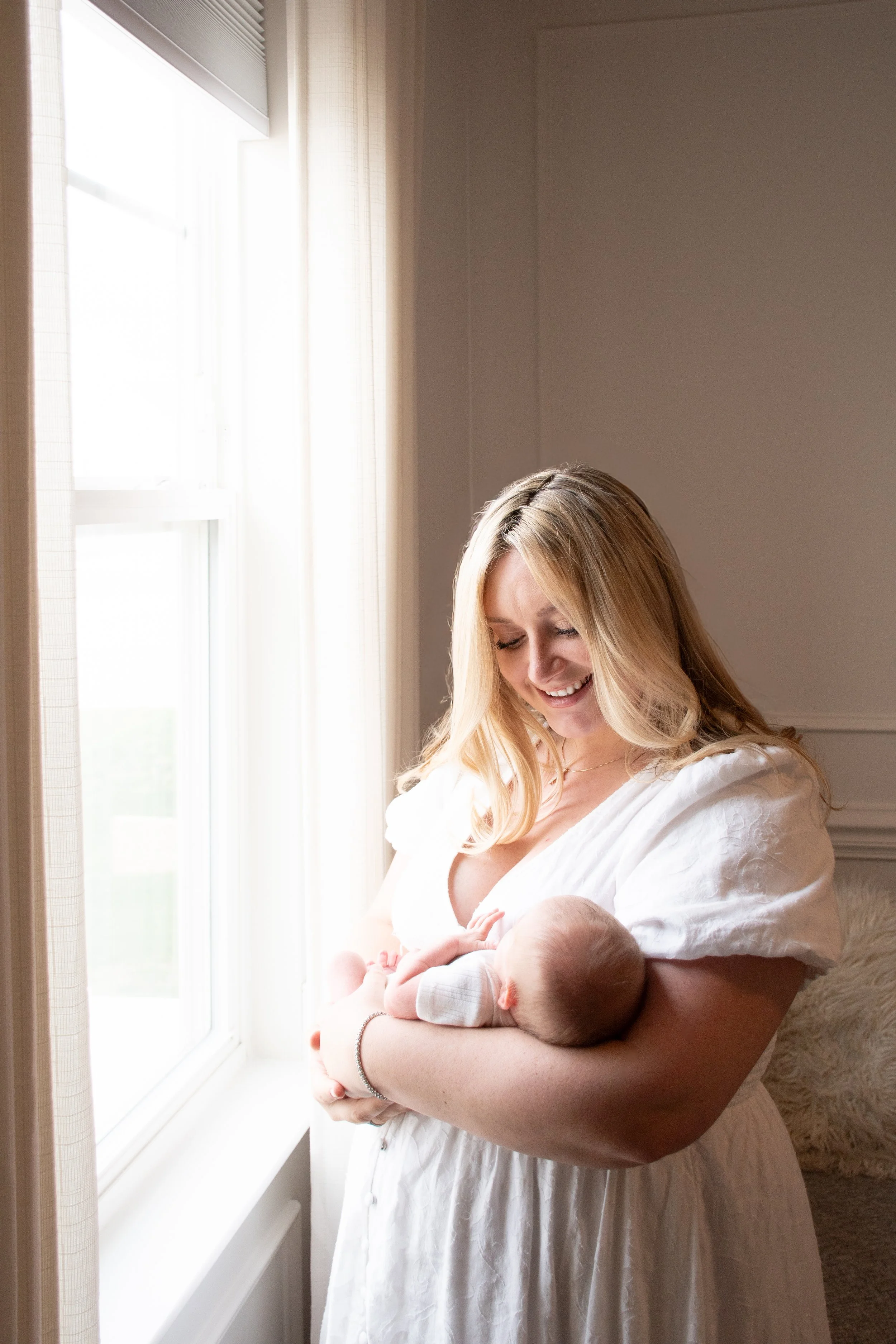 A woman in a white dress smiling as she holds her baby by a bright window.