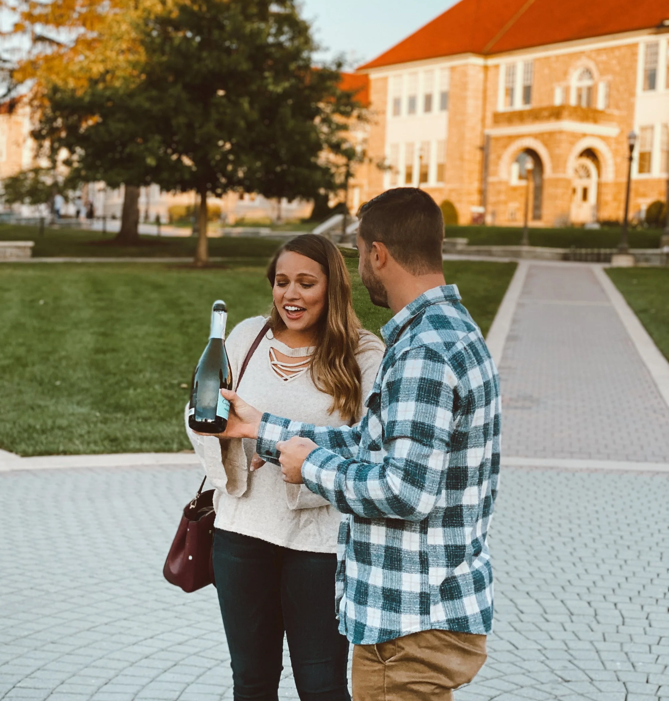 A man and woman standing outside on a paved walkway in a park, looking at a bottle of champagne together, with a large brick building and trees in the background during sunset.
