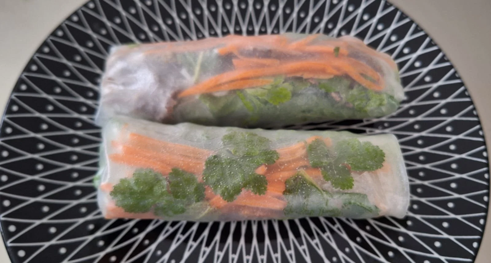 Deux rouleaux de printemps frais avec des légumes, tels que carottes, coriandre et vermicelles, emballés dans une feuille de riz transparente, placés sur une assiette à motif noir et blanc.
