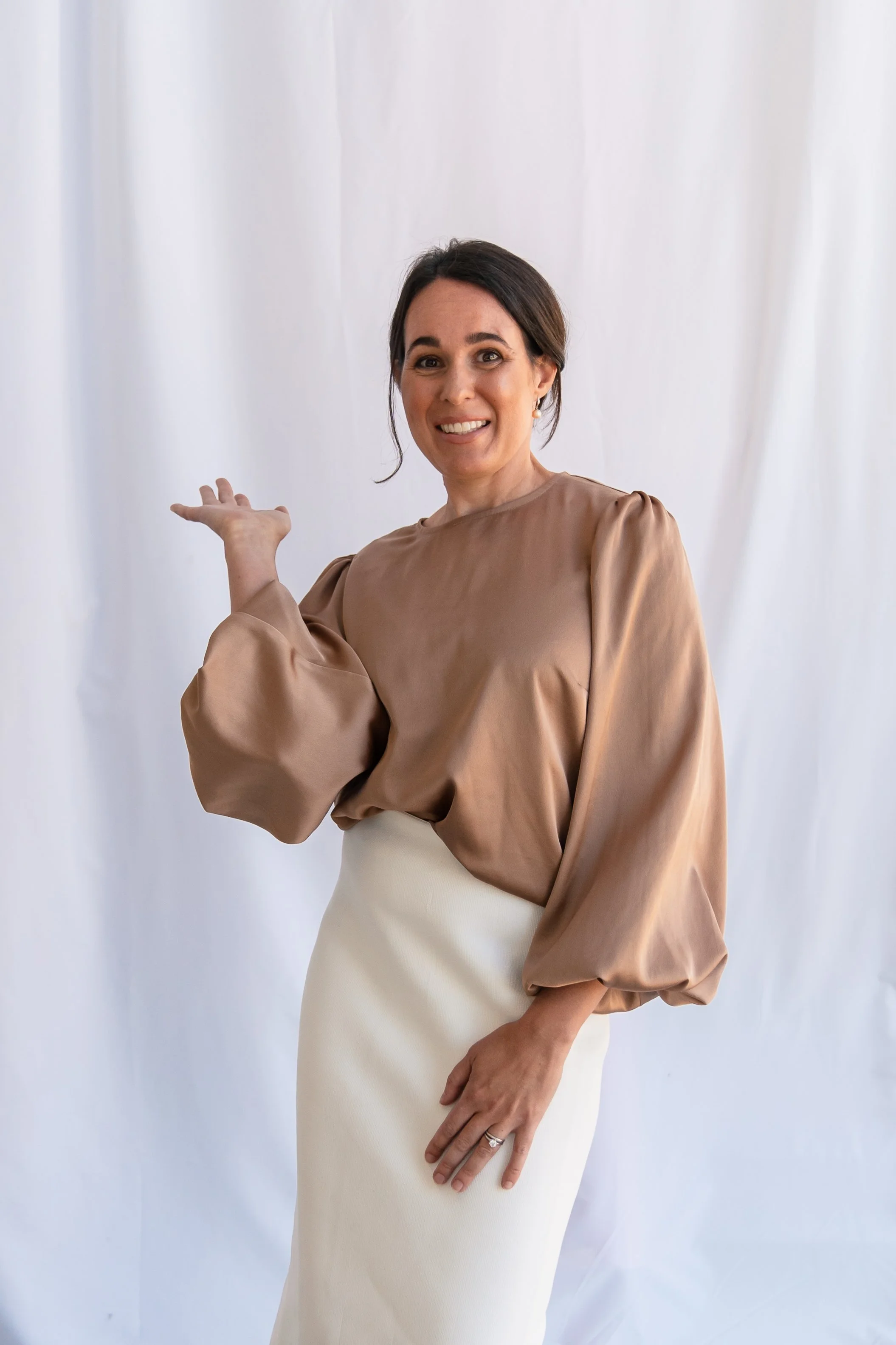 A woman with dark hair and earrings smiling, wearing a tan blouse with puffy sleeves and a white skirt, pointing to the side against a white background.