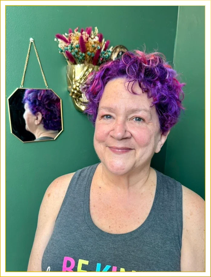 Woman with short, curly, purple hair, smiling, wearing a gray tank top, in a room with green walls, decorated with a mirror, floral arrangements, and a decorative wall hanging.