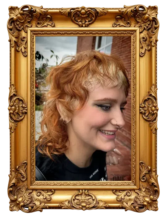 A young woman with curly red hair, smiling, seen through an ornate gold picture frame, with a brick building and windows in the background.