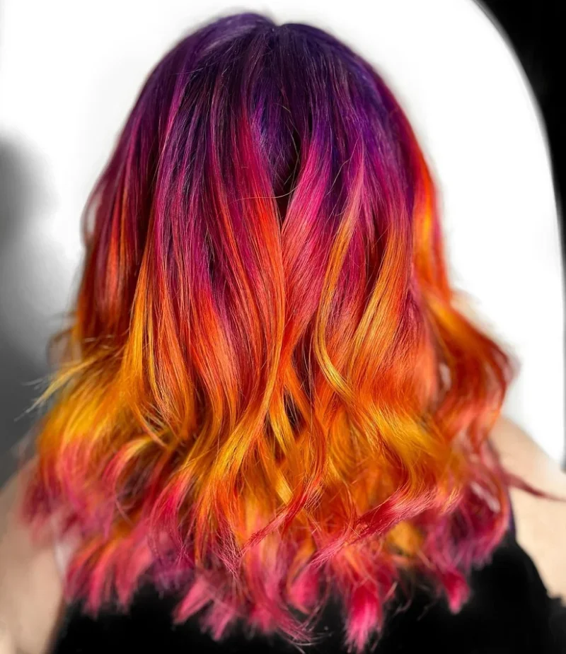 Back of a woman's head with shoulder-length wavy hair dyed in a multi-color gradient, including purple at the top blending into pink, orange, and yellow towards the ends.