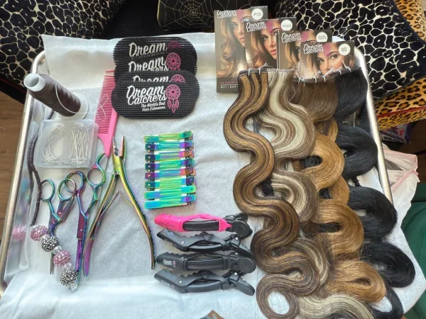 A tray displaying hairdressing supplies including hair clips, hair extension hairpieces, a comb, scissors, curlers, and hair dye bottles.