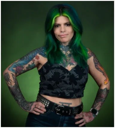 Woman with green and blue hair, tattoos on arms and torso, wearing a black crop top and belt, standing with hands on hips against a green background.