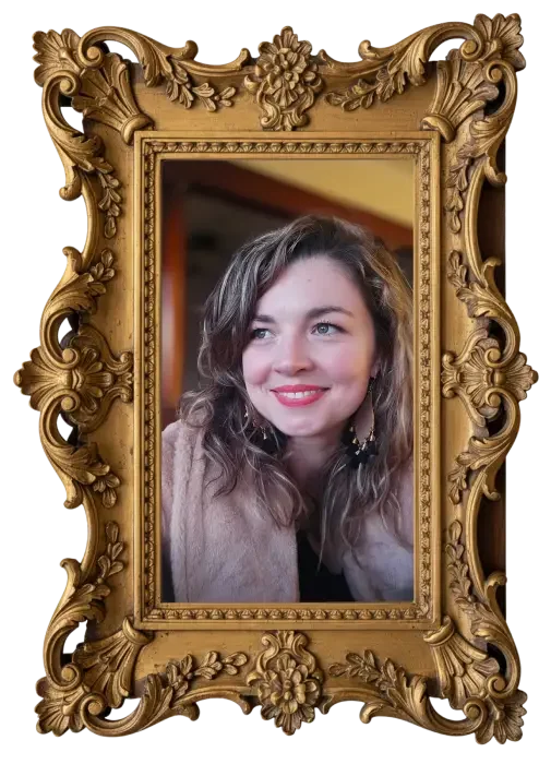 A woman with wavy light brown hair and blue eyes, smiling, is posing for a photo inside an ornate, gold-colored picture frame.