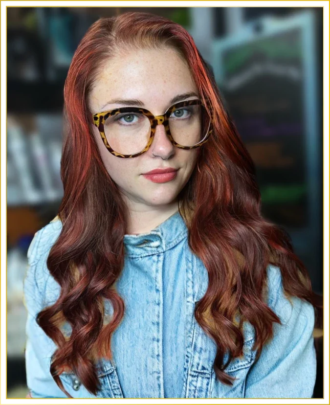 Young woman with red wavy hair wearing large tortoise shell glasses and a denim jacket, looking at the camera.