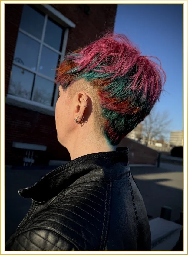 A person with a multi-colored, edgy hairstyle with pink, green, and orange hues, wearing a black leather jacket and multiple earrings, standing outdoors in front of a building with a window.