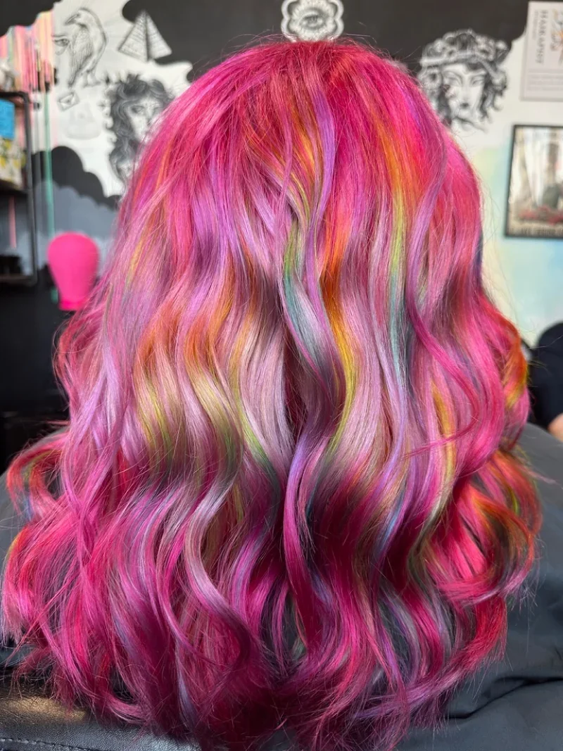 Back view of a person with long, wavy hair dyed in multiple bright colors including pink, purple, orange, green, and blue.