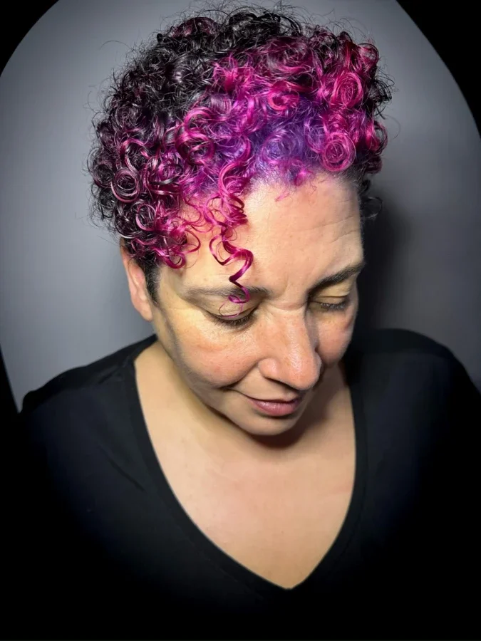 Close-up of a person's hair with multicolored rainbow shades including blue, red, yellow, and green, and wavy texture, outdoors with brick wall background.