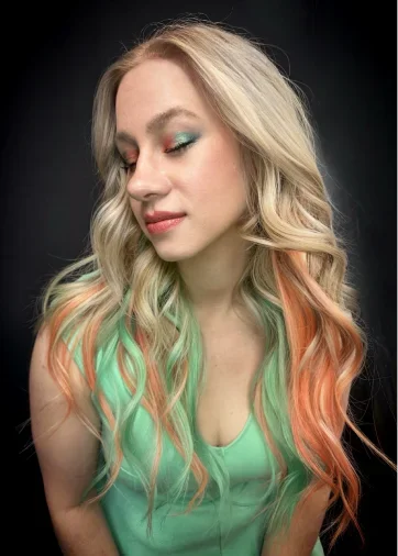 A woman with platinum blonde hair featuring colorful pastel pink and green streaks, wearing a mint green top and vibrant makeup with teal eyeshadow.
