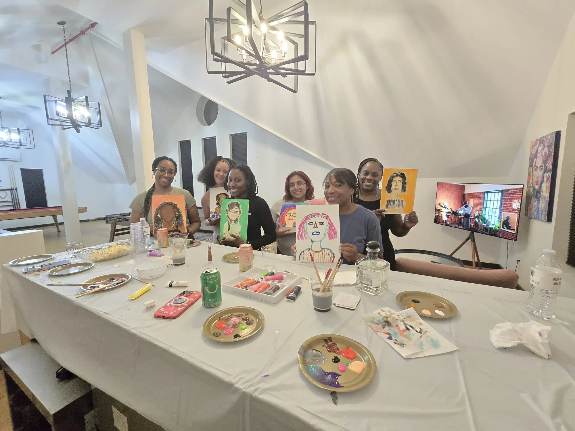 Group of women participating in a painting activity at a table, displaying their artwork, with art supplies, food, and a television in a brightly lit room.