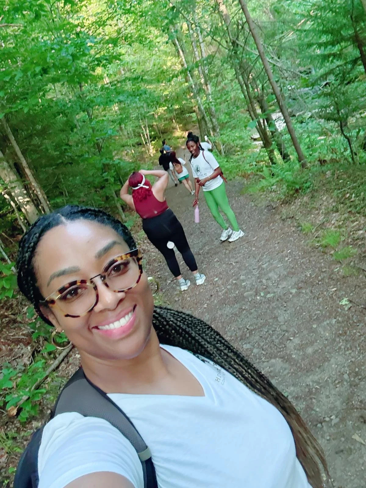 A woman with braided hair and glasses taking a selfie on a wooded trail, with several people walking in the background.