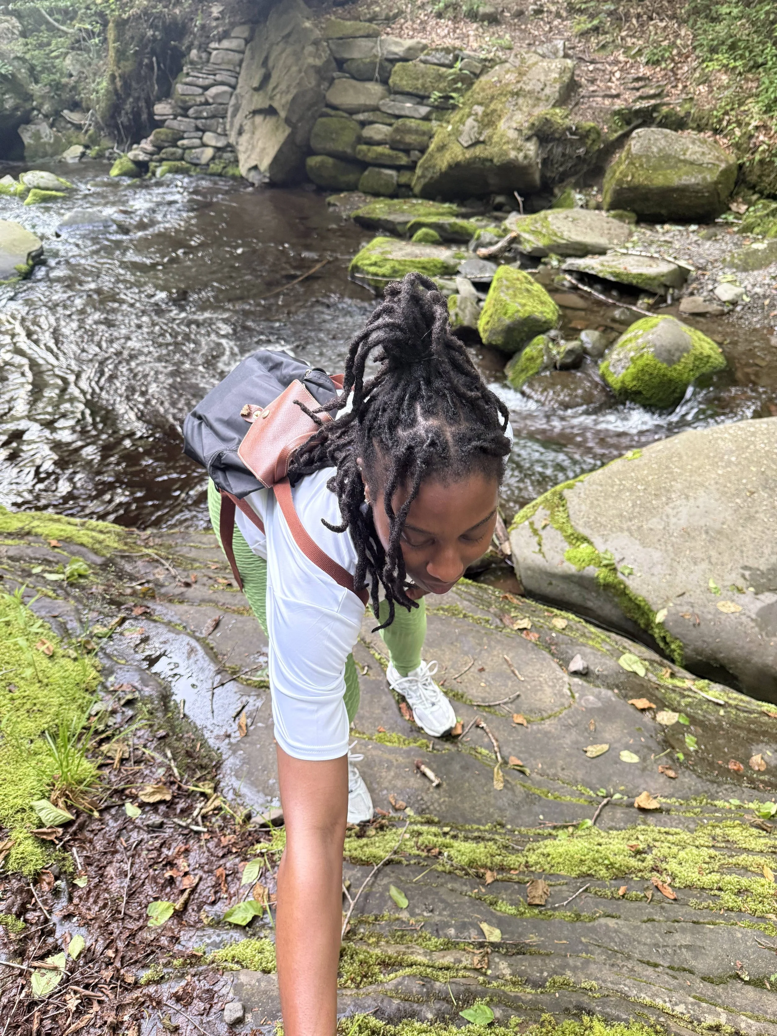 A person with long dreadlocks, white sports shirt, green pants, and white sneakers climbing on mossy rocks beside a stream in a forest.