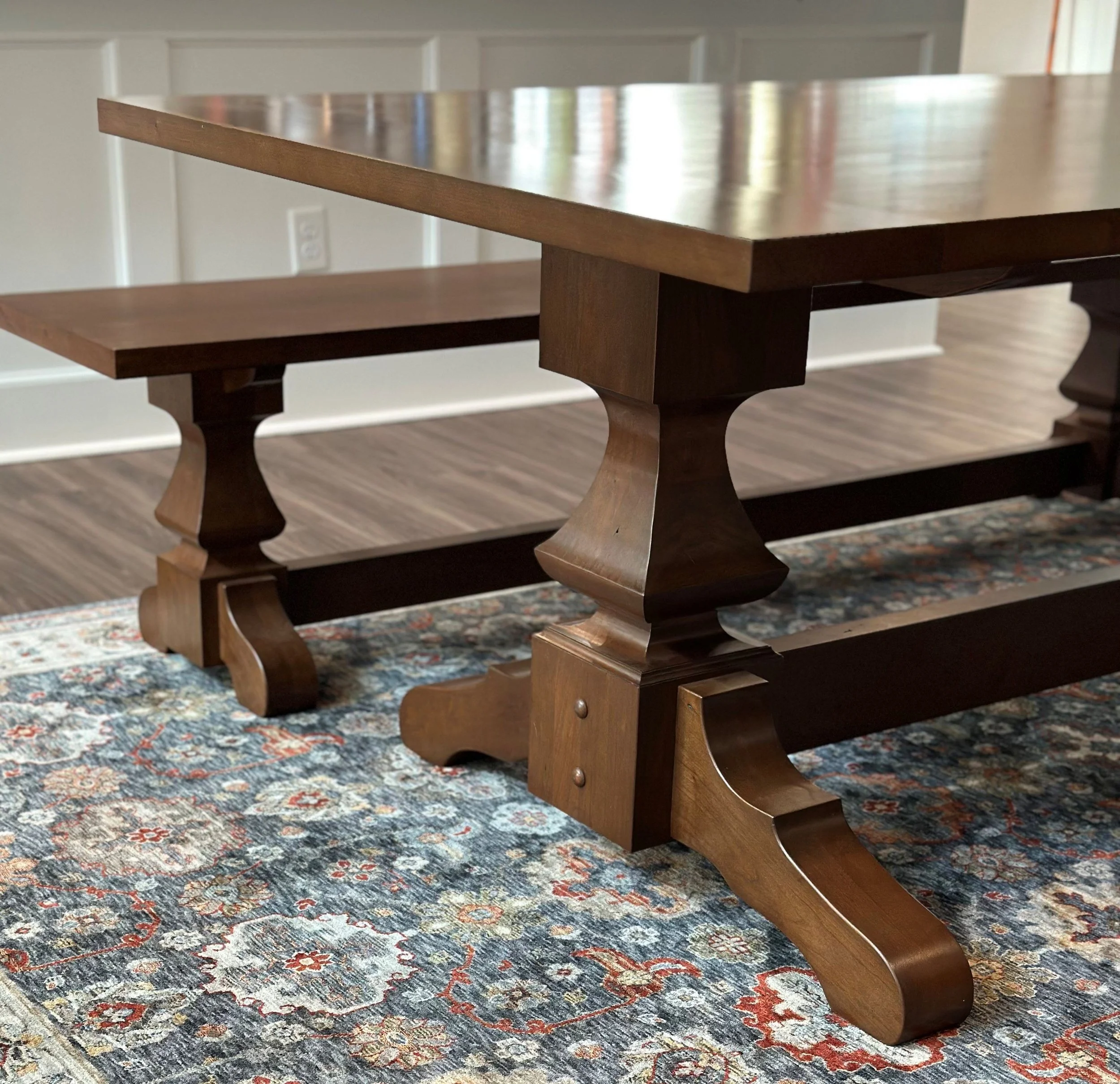 Close-up of a wooden dining table with ornate legs, placed on a patterned area rug in a room with wooden flooring and white walls.