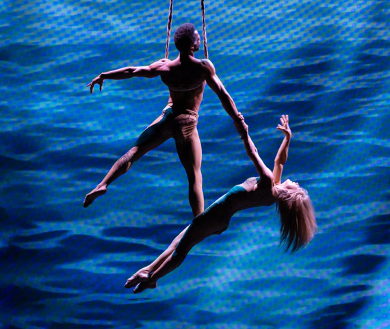 Two acrobatic performers, one male and one female, suspended over water, holding hands as the female hangs upside down.