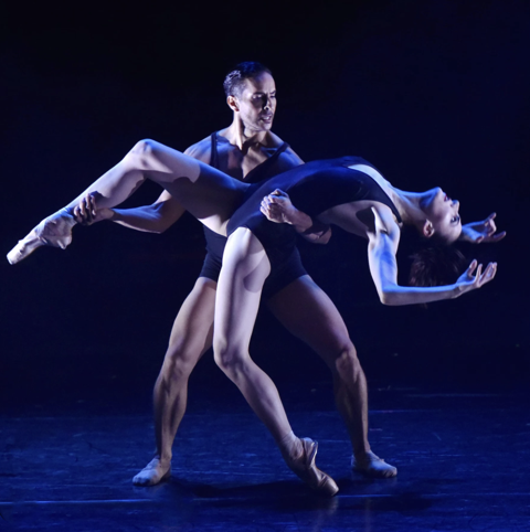 Two ballet dancers perform on stage with a dark background, one lifting the other in a dance pose.
