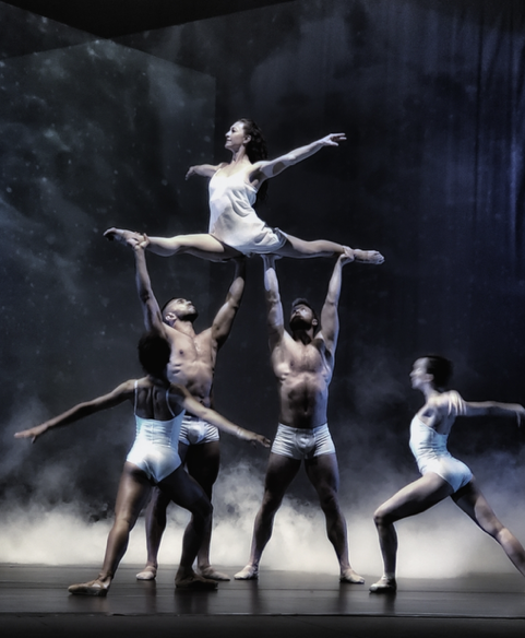 Ballet dancers performing on stage, with a female dancer balanced on the hands of male dancers, surrounded by a misty background.