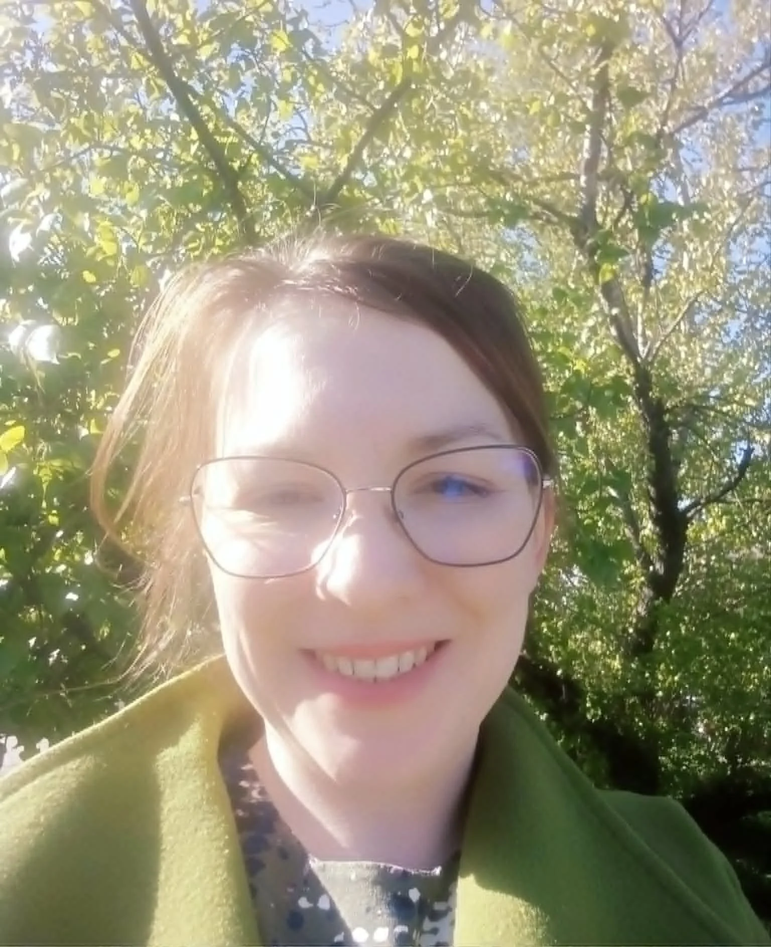 A smiling person with glasses and brown hair, outdoors under trees with yellow and green leaves, taking a selfie.