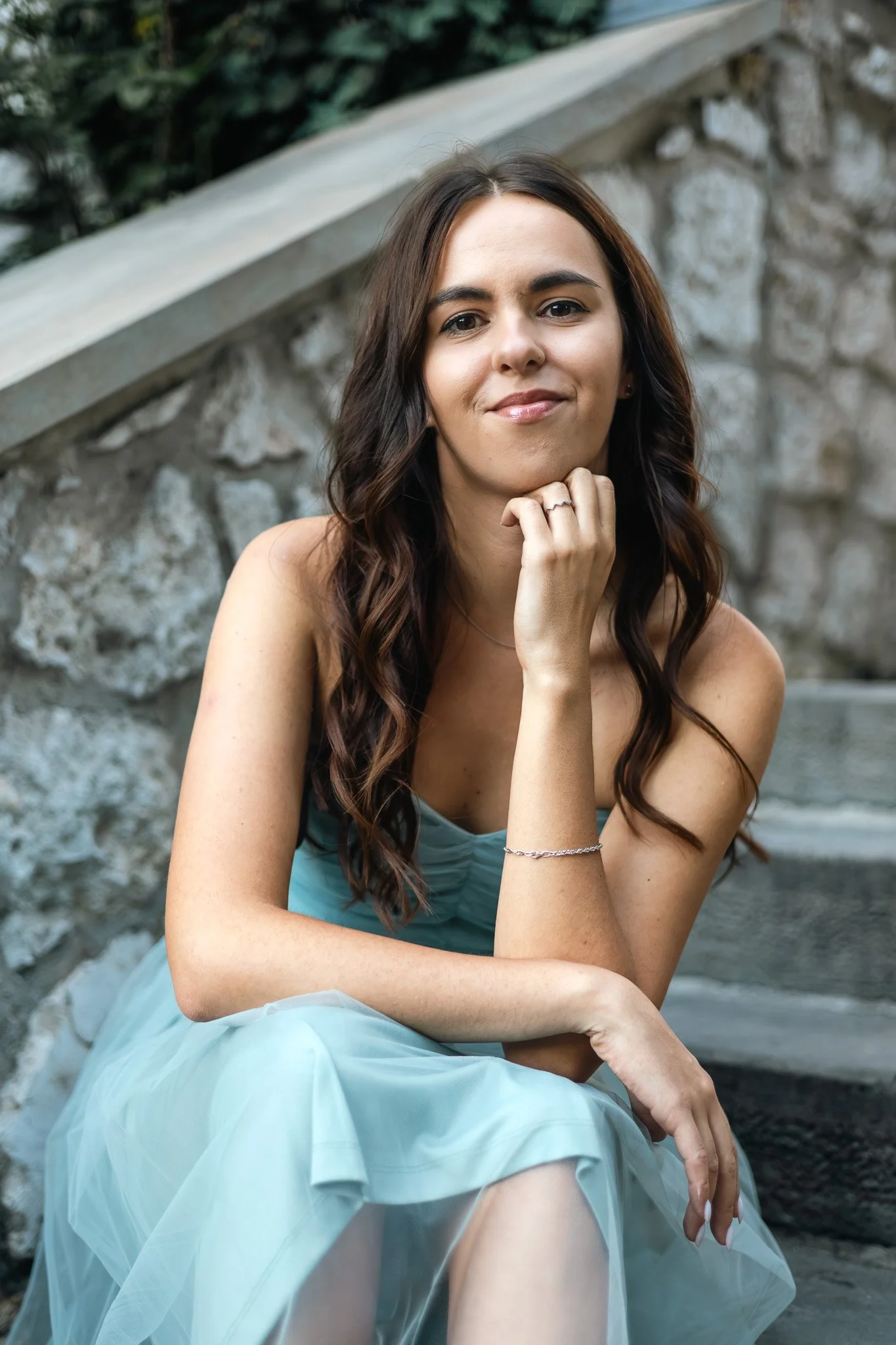 A woman with long, wavy brown hair, sitting outdoors near a stone wall and metal railing, wearing a light blue, sleeveless dress, smiling gently, resting her chin on her hand.