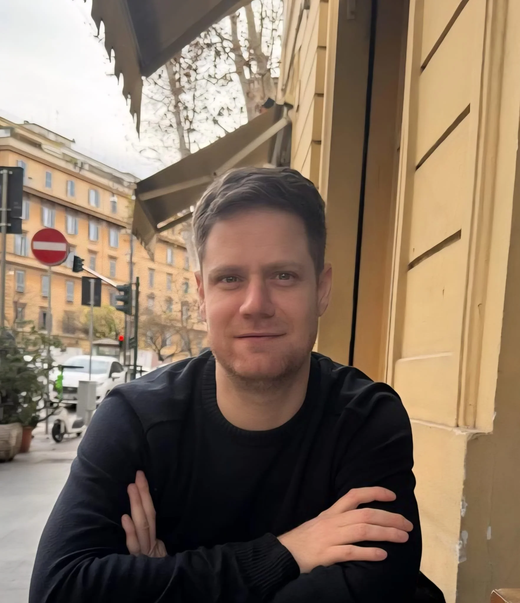 A man with short hair and a friendly expression, wearing a black jacket, is standing outside in front of a yellow building with window shutters. He is crossing his arms and looking at the camera. There are trees, a street with cars, and a no-entry traffic sign visible in the background.