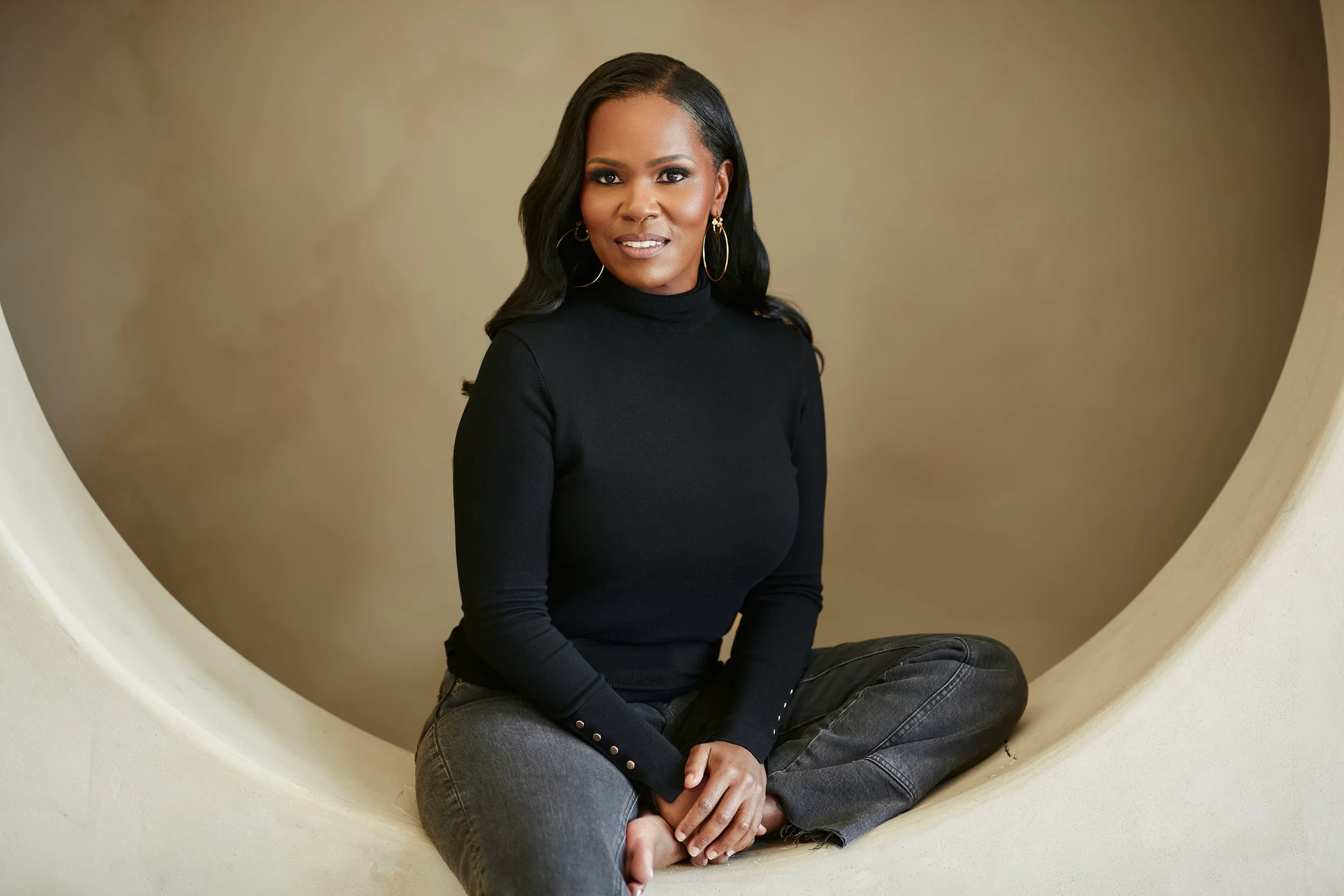 A woman sitting cross-legged inside a circular opening, wearing a black turtleneck and gray jeans, with long dark hair and hoop earrings, looking at the camera.