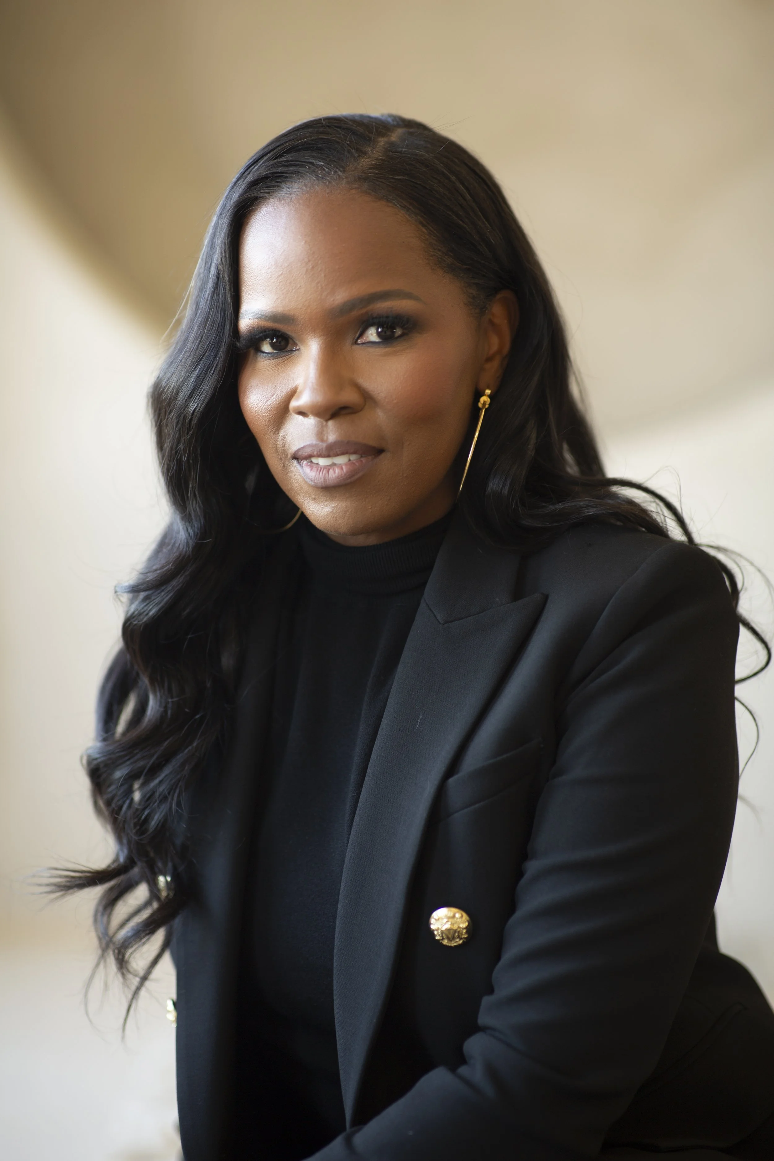 Professional woman with long dark hair wearing a black blazer and gold jewelry, looking at the camera.
