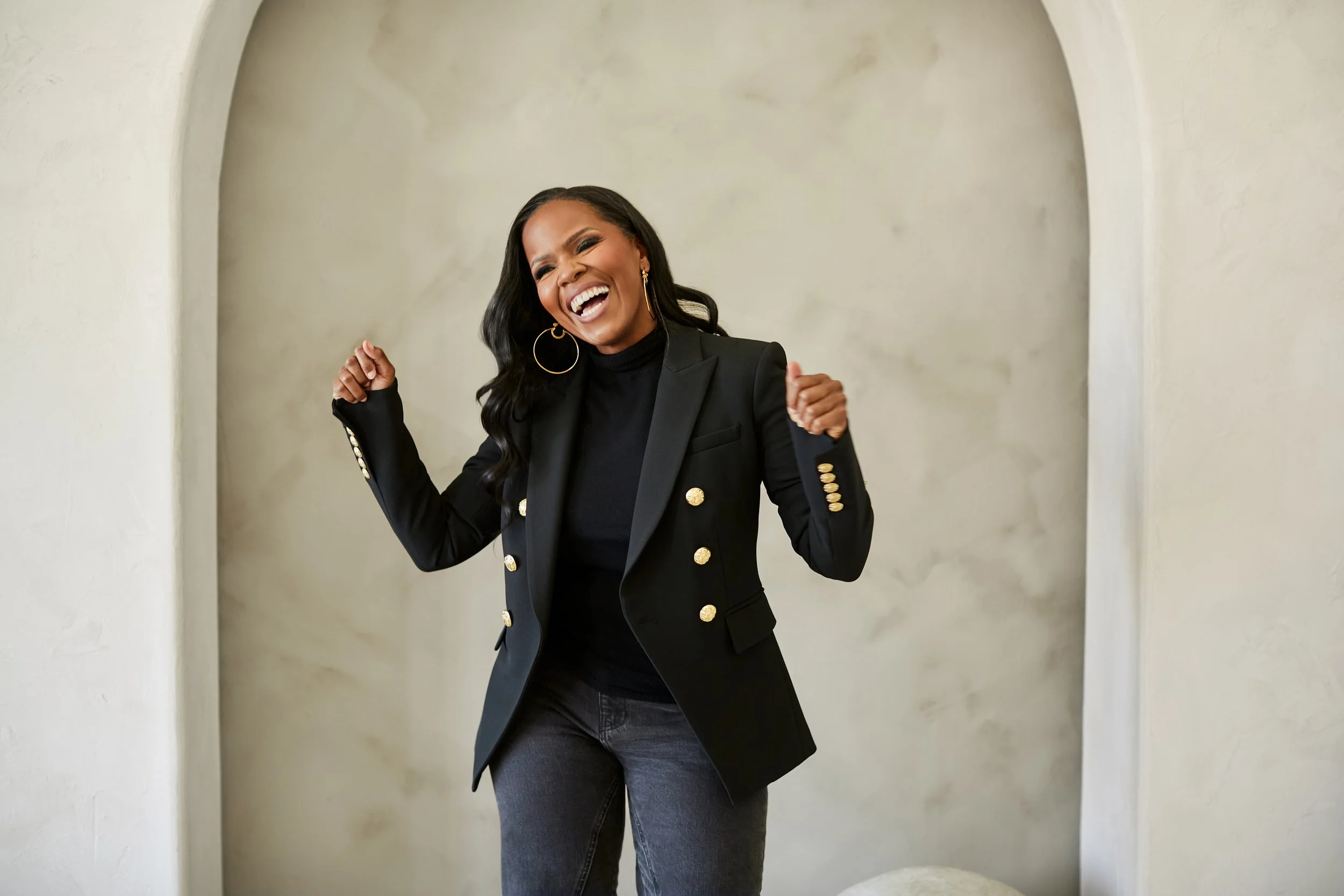 A woman with long black hair, wearing a black blazer with gold buttons, black top, and black pants, is smiling and celebrating with fists raised in an indoor setting.